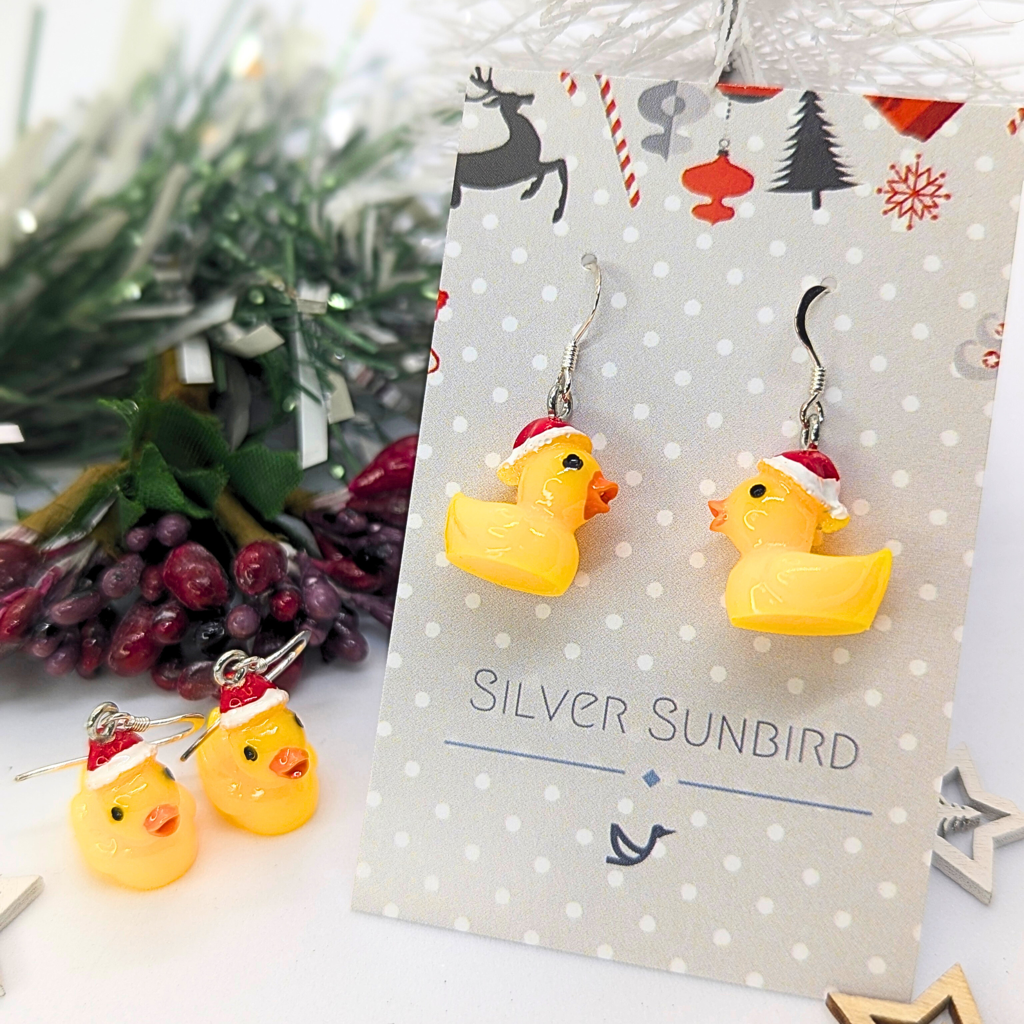 Festive Duck Earrings by Silver Sunbird, showcasing yellow rubber ducks adorned with Santa hats, are presented on a Silver Sunbird card. In the background, you can see holiday-themed decorations and coordinating stud earrings.