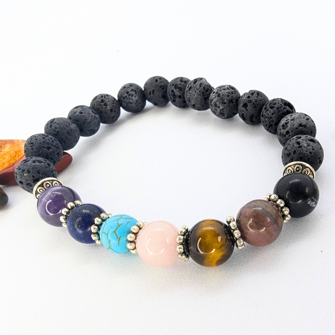 The Chakra Bracelet by Silver Sunbird features black lava stones and seven colorful semi-precious gemstones, separated by silver spacers. Use the lava stones with essential oils for extra benefits. Displayed on a white background.