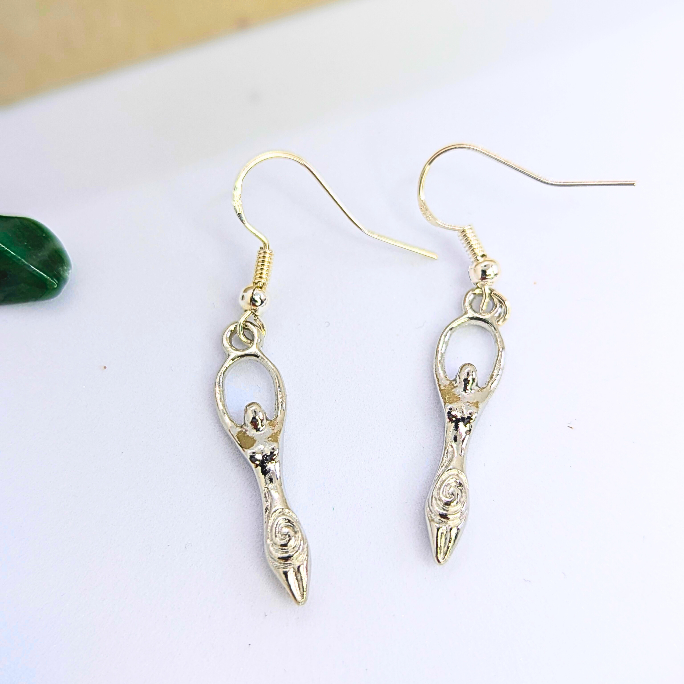 Silver Sunbird’s Fertility Goddess Dangle Earrings feature silver-tone abstract figures with uplifted arms and spiral accents, displayed on a white surface—an eye-catching piece of symbolic jewellery.
