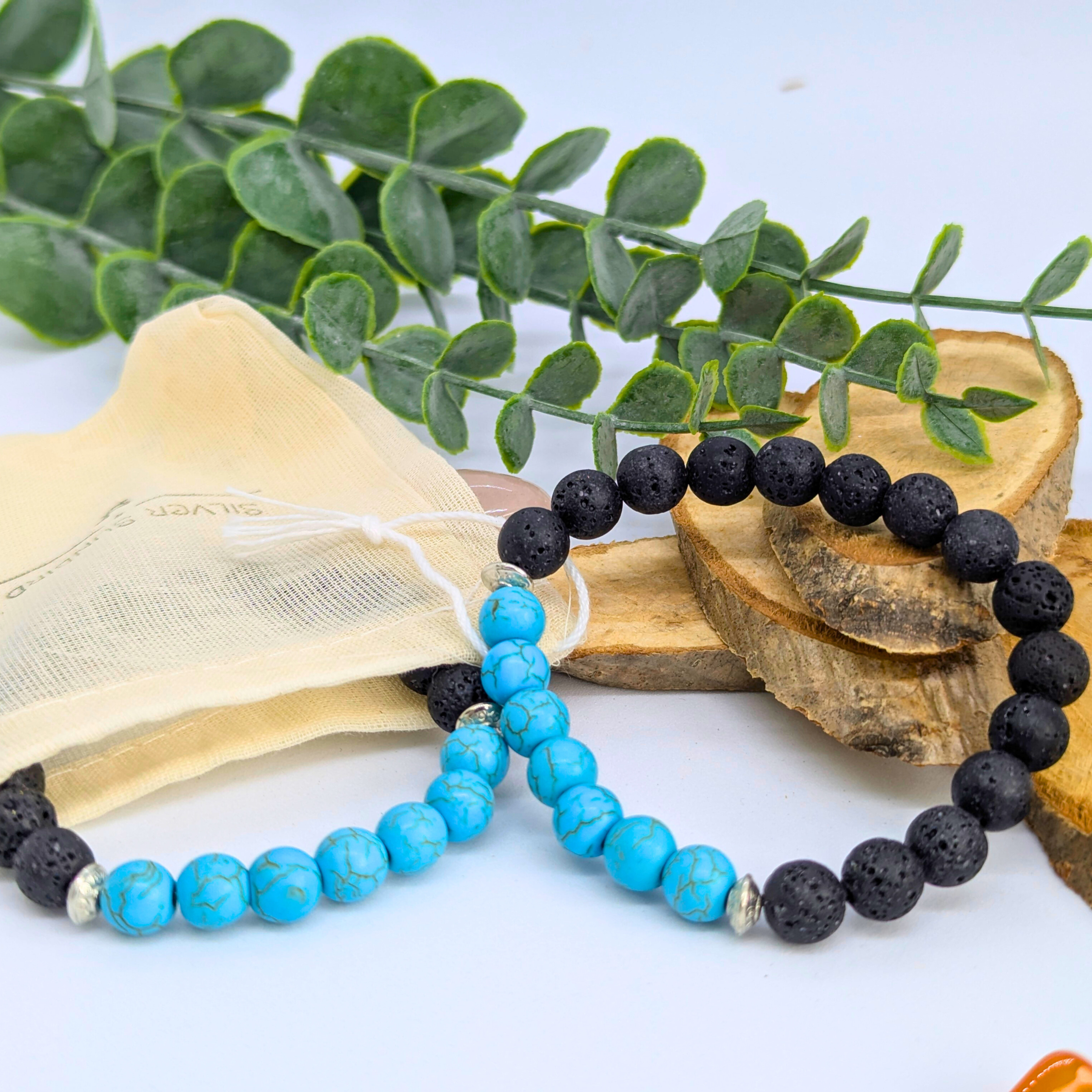 Two Silver Sunbird Turquoise and Lava Stone Bracelets are shown on wooden pieces, with green leaves and a beige drawstring pouch in the background. One bracelet features turquoise and black beads; the other is mostly black.