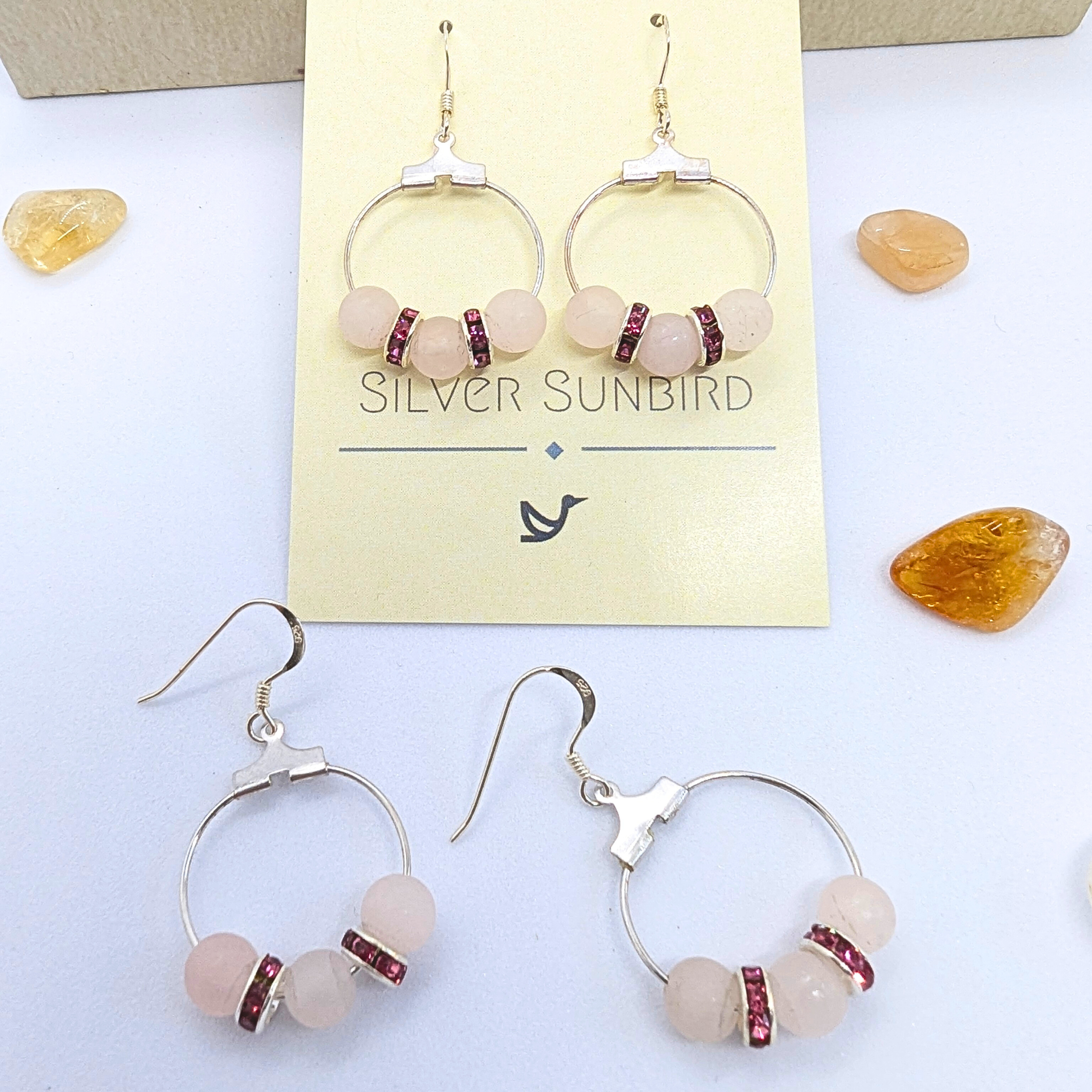 A pair of Rose Quartz Statement Earrings from Silver Sunbird displayed on a card labeled "Silver Sunbird." The earrings feature circular silver hoops adorned with pink gemstones and small red rhinestone spacers. Four additional gemstones are scattered on a white surface around the card.