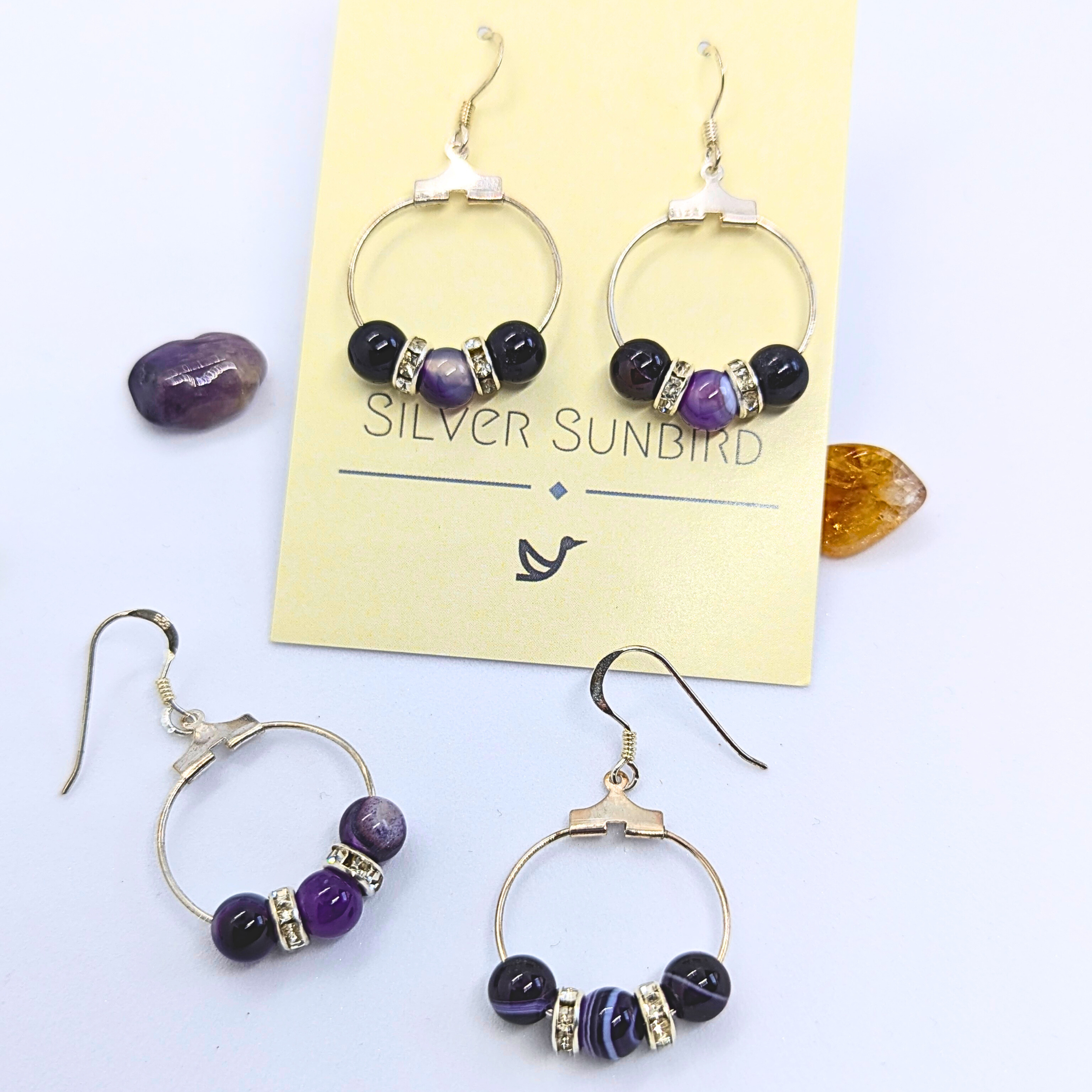 A pair of stylish Amethyst Statement Large Earrings lay on a card labelled "Silver Sunbird." The sterling silver earrings are circular with purple amethyst round gemstones, adorned with small silver rhinestone spacers. Beside the card are two loose matching earrings and three natural gemstones in shades of purple and amber.