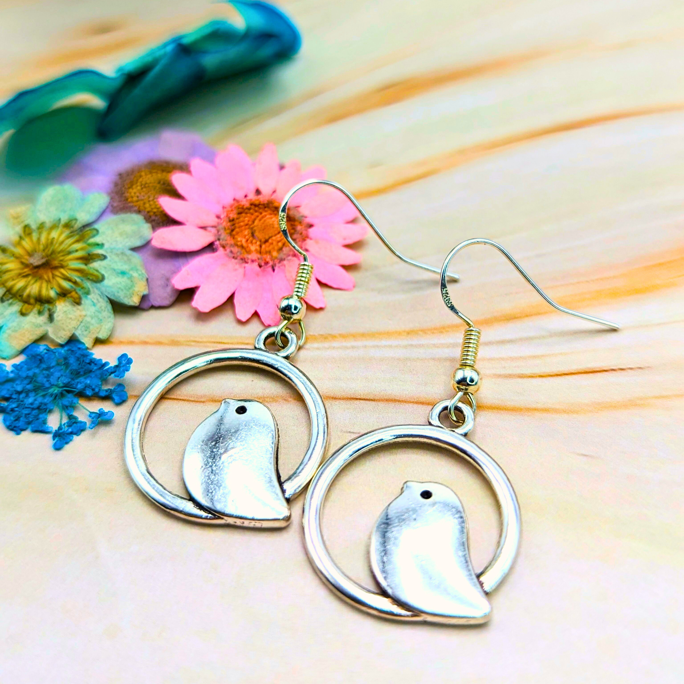 Beautiful Bird Earrings by Silver Sunbird, made from 925 Sterling Silver, rest on a wooden surface with colorful dried flowers and greenery. Perfect for bird lovers, these earrings feature small birds within circular frames, adding a touch of nature-inspired elegance.