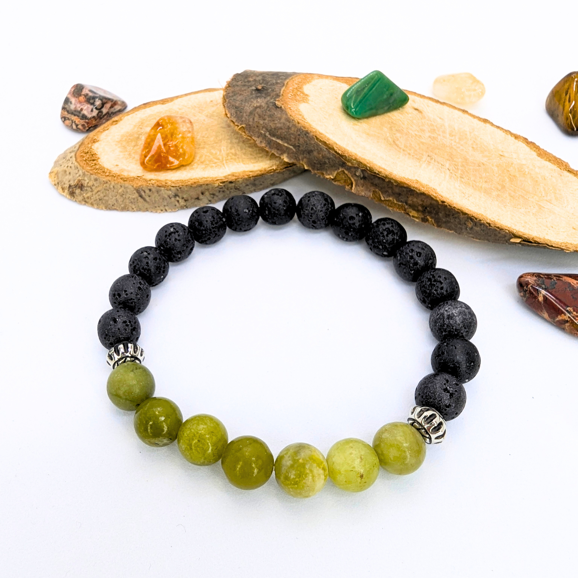 The Silver Sunbird Peridot and Lava Stone Bracelets, featuring green and black gemstone beads with silver accents, are displayed on a white surface with sliced wood pieces and colorful tumbled stones.