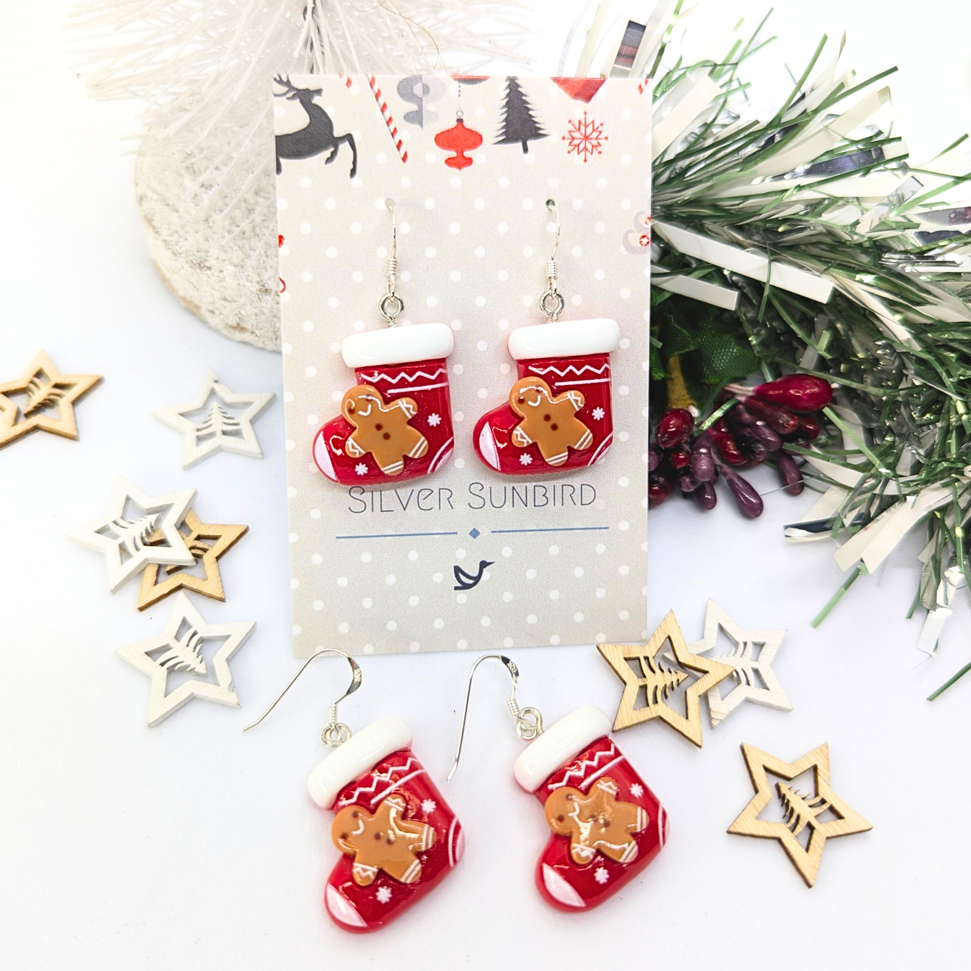Gingerbread Men Stocking Earrings from Silver Sunbird dangle elegantly on a card, surrounded by decorative stars and greenery, making them the perfect charming holiday accessories to add a touch of cheer.