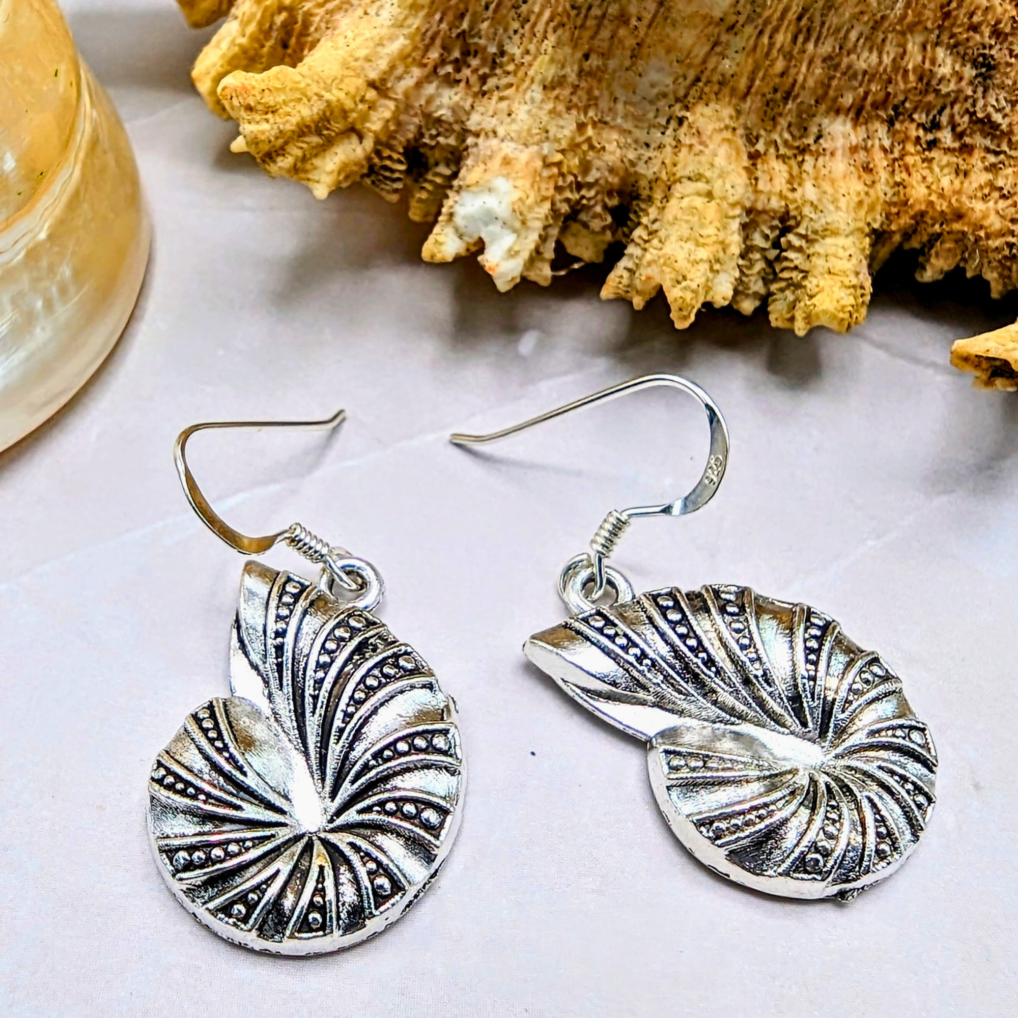 The Shoreline Spiral Shell Earrings by Silver Sunbird, featuring intricate nautilus-inspired designs, rest beside a textured seashell—ideal for lovers of ocean-inspired jewellery.