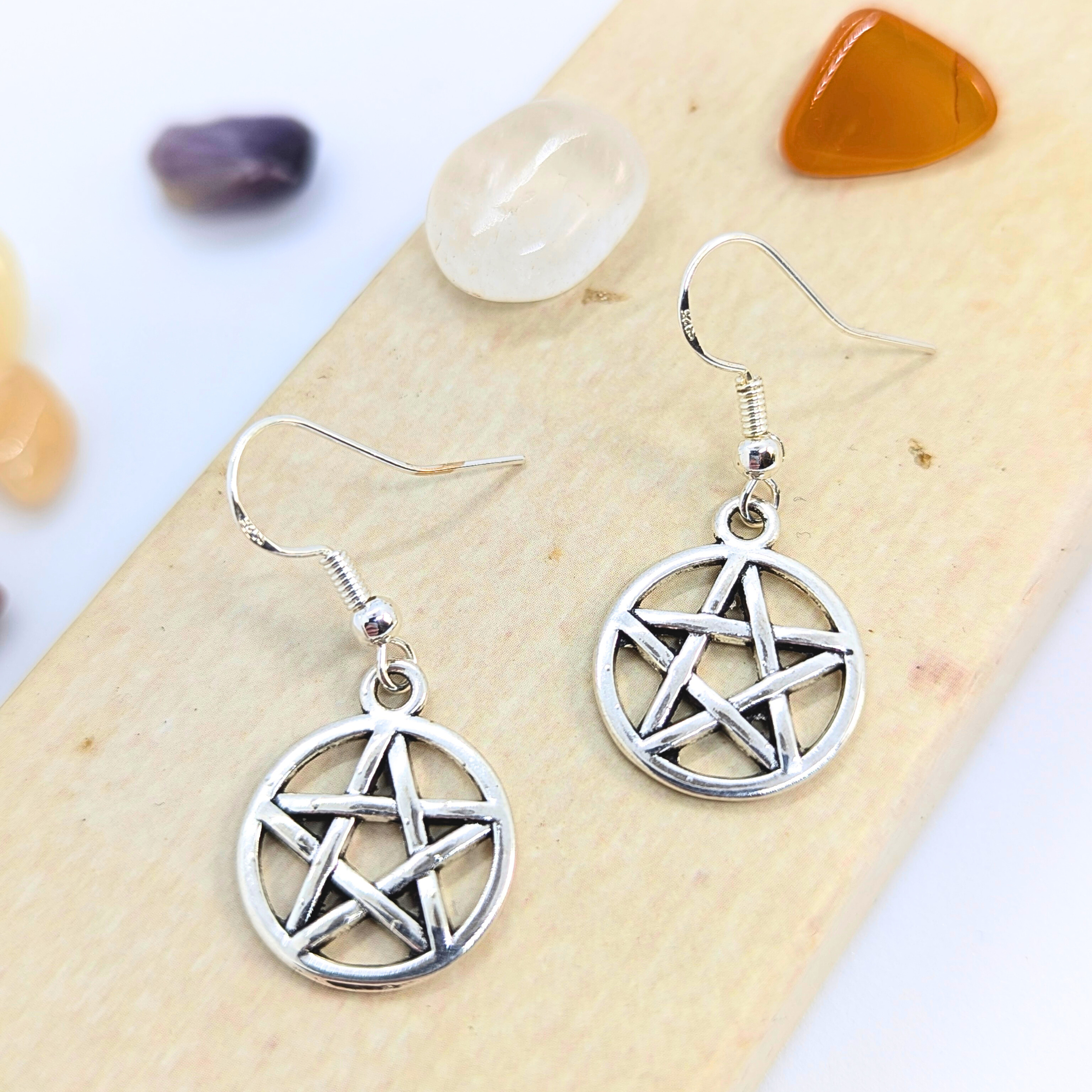 Silver Sunbird Pentagram Earrings are displayed on a light surface, encircled by polished white, orange, and purple stones—ideal for fans of Wicca jewelry or pagan symbolism.