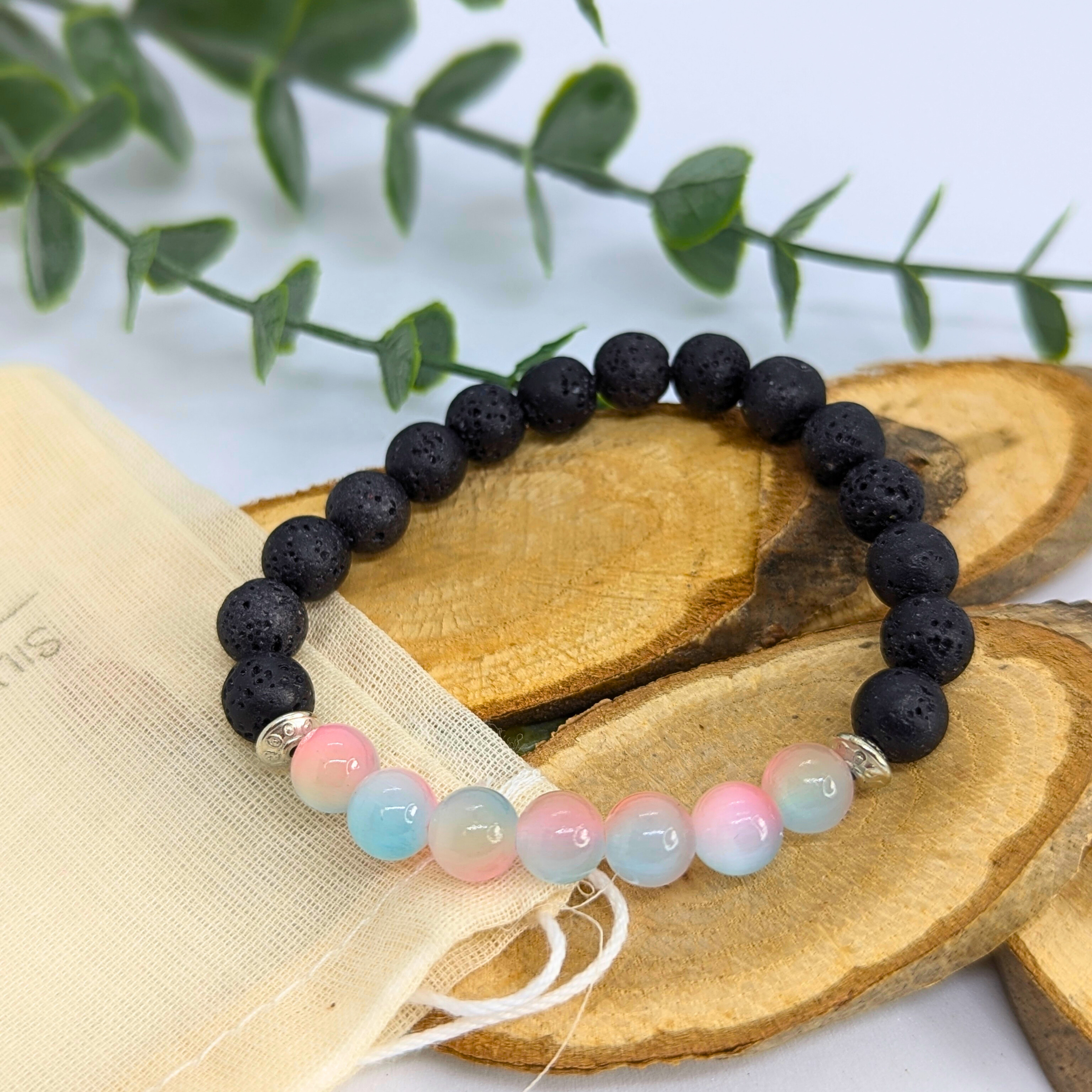 The Silver Sunbird Mermaid-Inspired Lava Stone Bracelet, featuring black lava stones and pastel blue and pink beads, is showcased on wooden slices beside a cream drawstring pouch—an ideal accessory for ocean lovers.