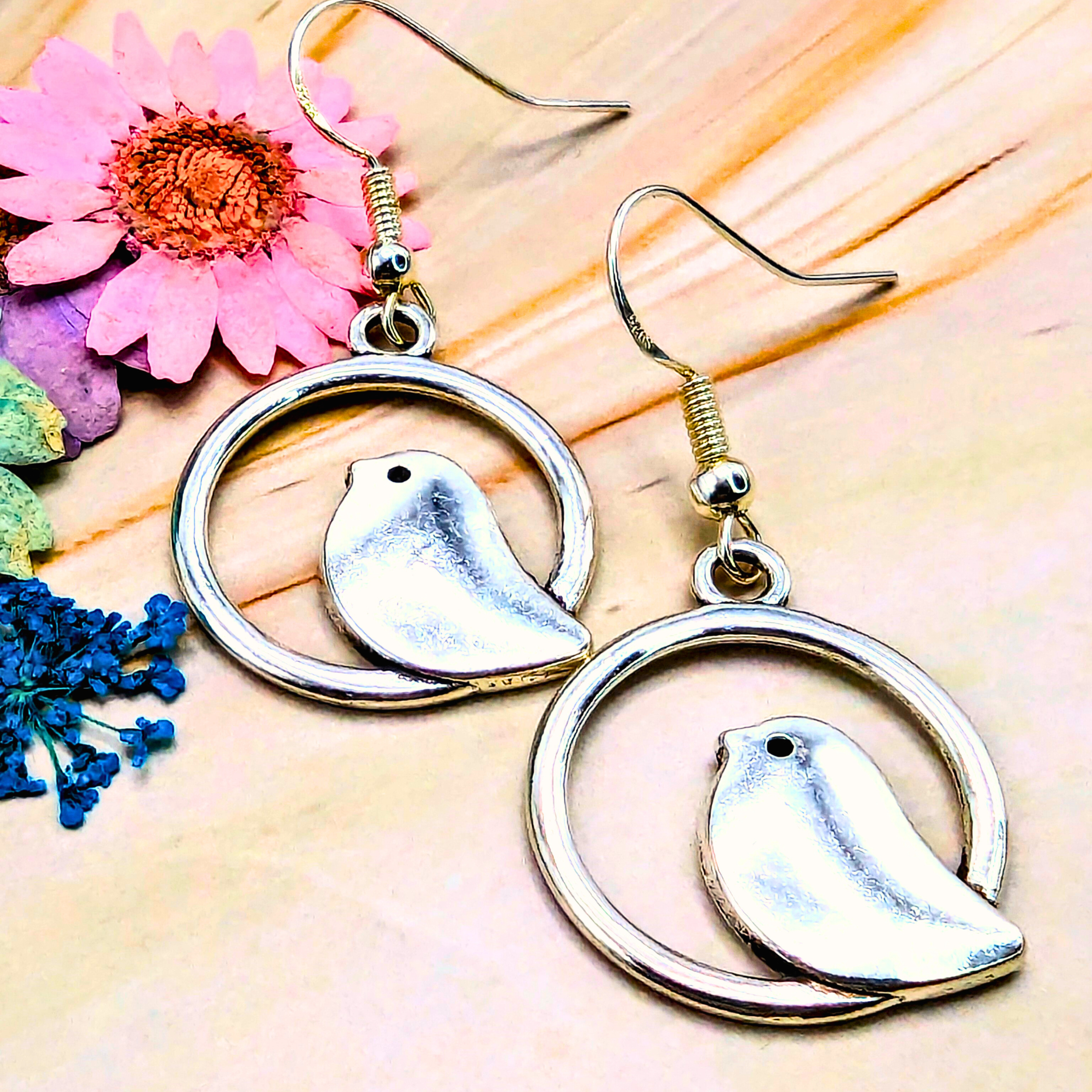 The Beautiful Bird Earrings by Silver Sunbird feature exquisite 925 Sterling Silver bird pendants in circular frames. Ideal for bird lovers, they rest on a wooden surface adorned with pink and blue flowers, adding a touch of nature&