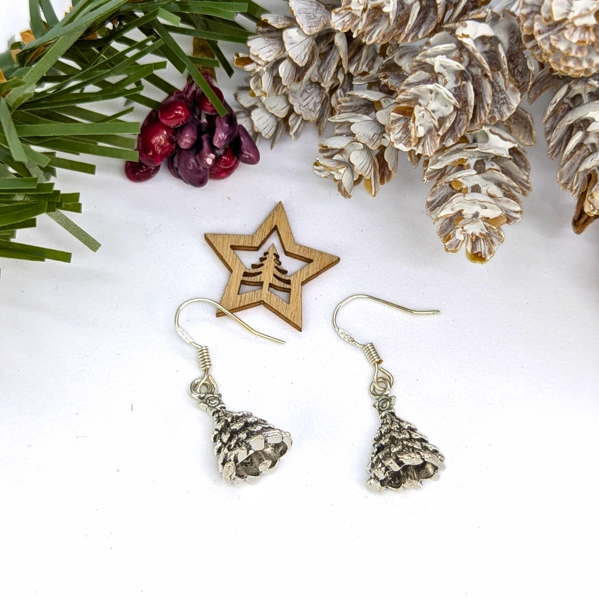 A pair of Silver Sunbird 3D Christmas Tree Earrings are displayed on a white surface among snow-dusted pinecones, evergreen branches, red berries, and a wooden star ornament.