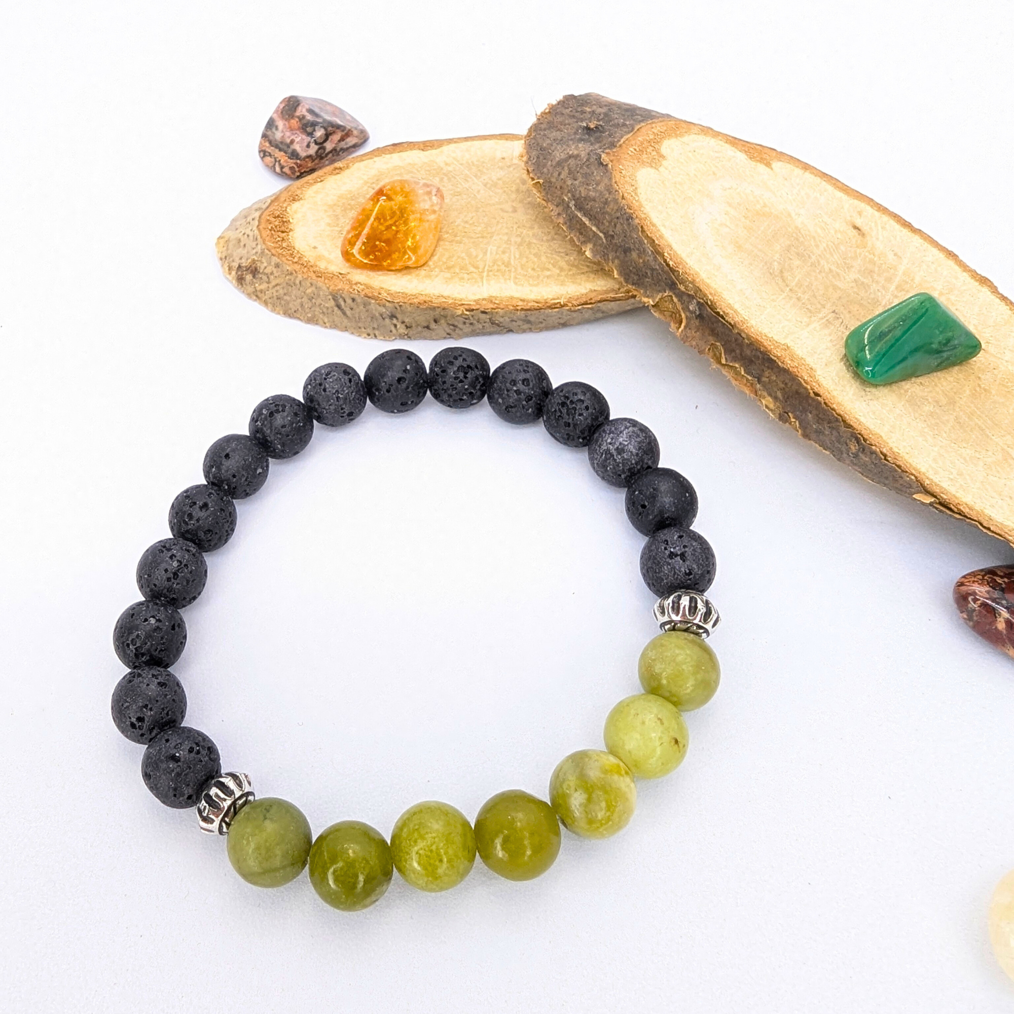 The Silver Sunbird Peridot and Lava Stone Bracelet, featuring black and green stones with silver spacers, is displayed on a white surface amid polished stones and wood accents.