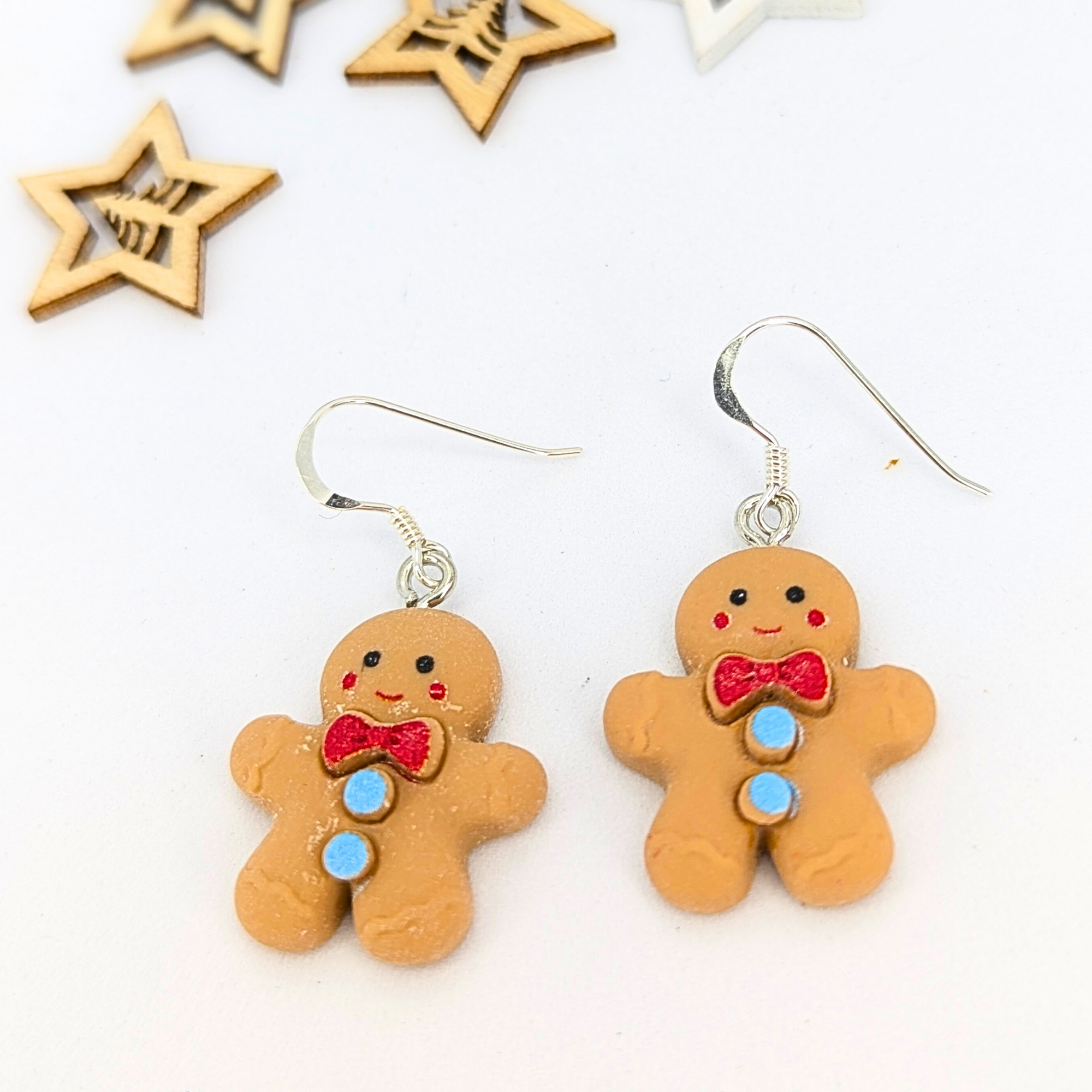 A pair of Cheerful Gingerbread Man Earrings by Silver Sunbird, featuring bright red bows and blue buttons, are adorned with silver hooks. They are showcased on a white surface with wooden star shapes in the background.