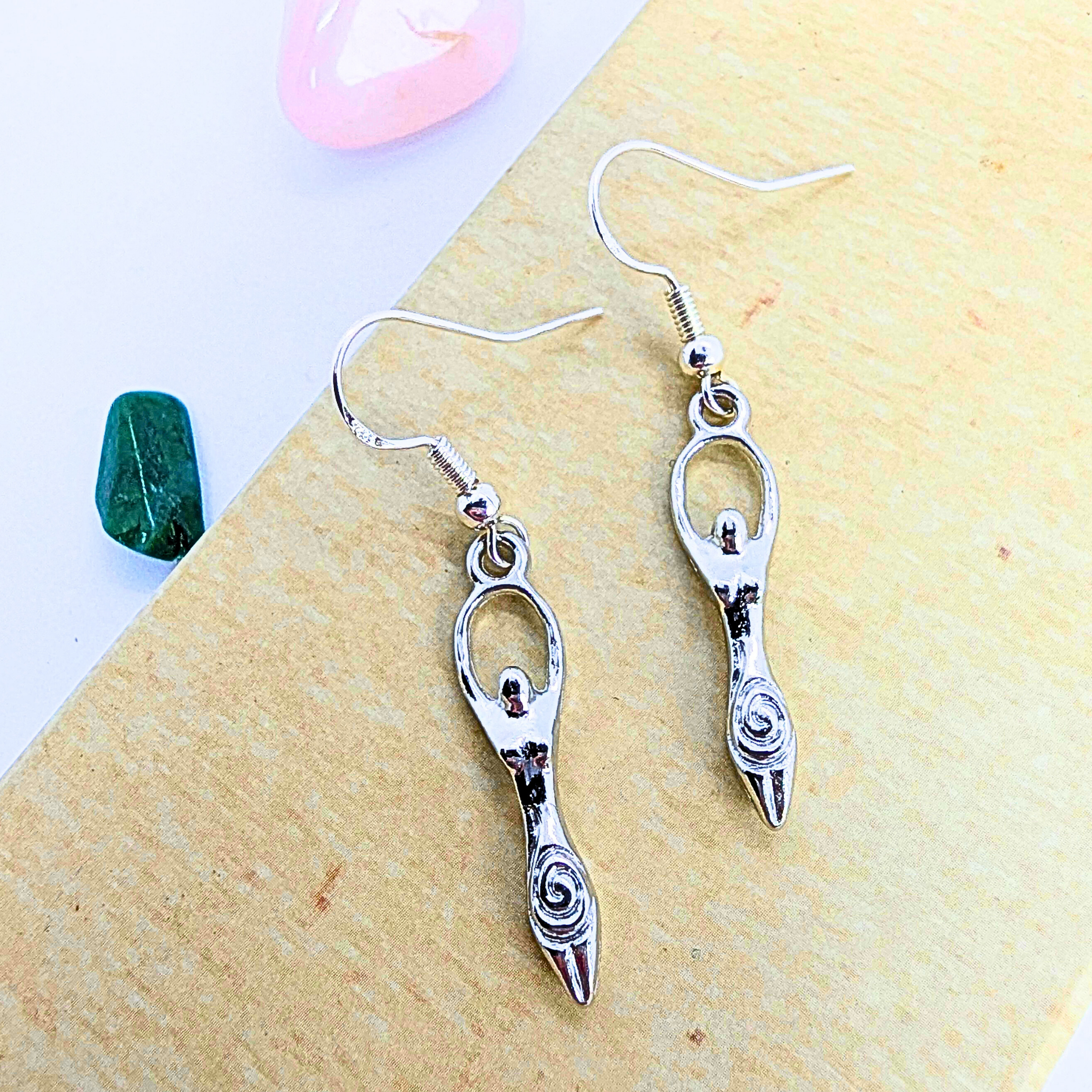 Silver Sunbird’s Fertility Goddess Dangle Earrings—sterling silver with spiral designs—are shown on a light wooden surface, next to a pink heart stone and green crystal, creating an elegant, symbolic jewellery display.
