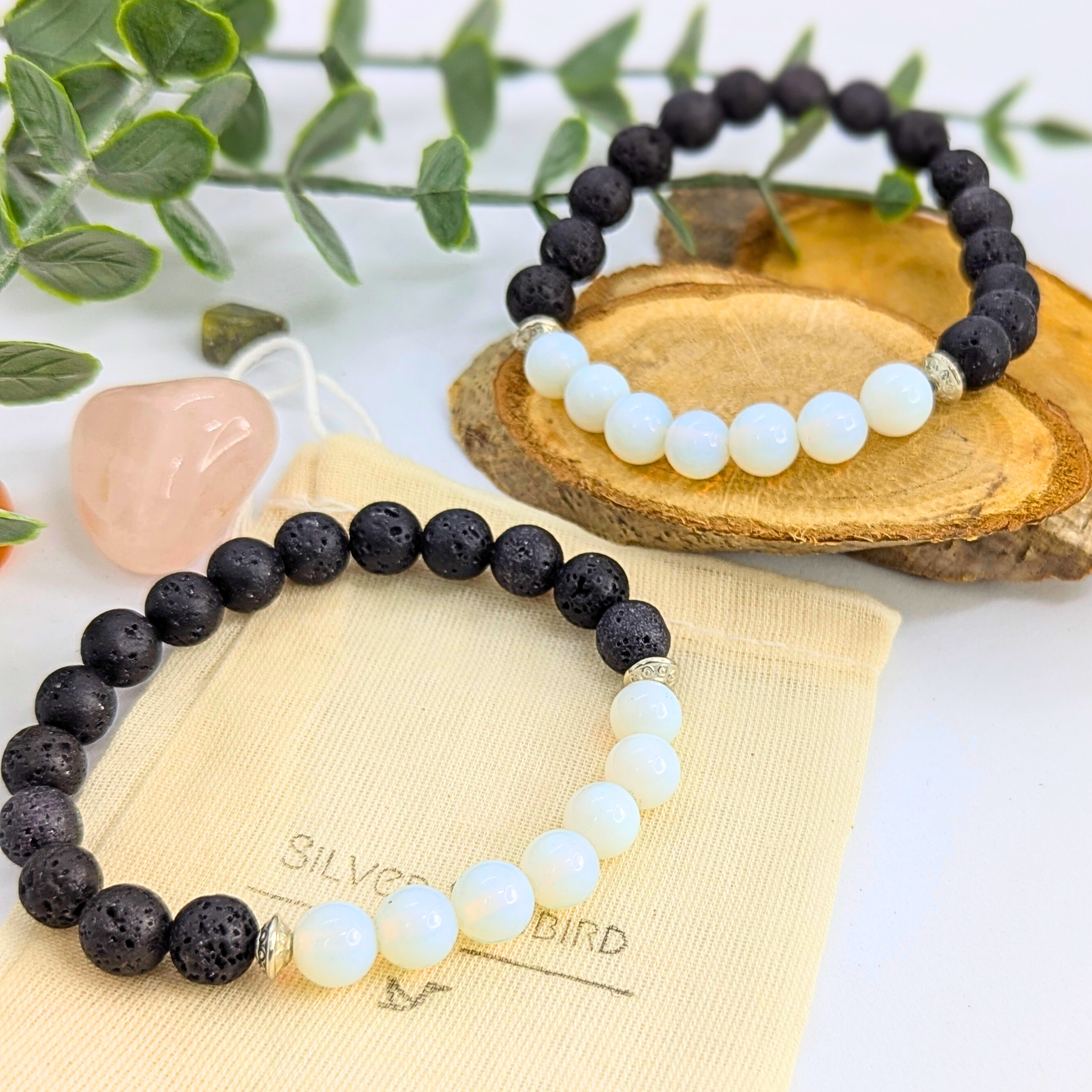 The Silver Sunbird Opal and Lava Stone Bracelets, featuring alternating black lava beads and white opal, are displayed on a cream pouch atop a wooden tray, surrounded by green leaves and a pink crystal.