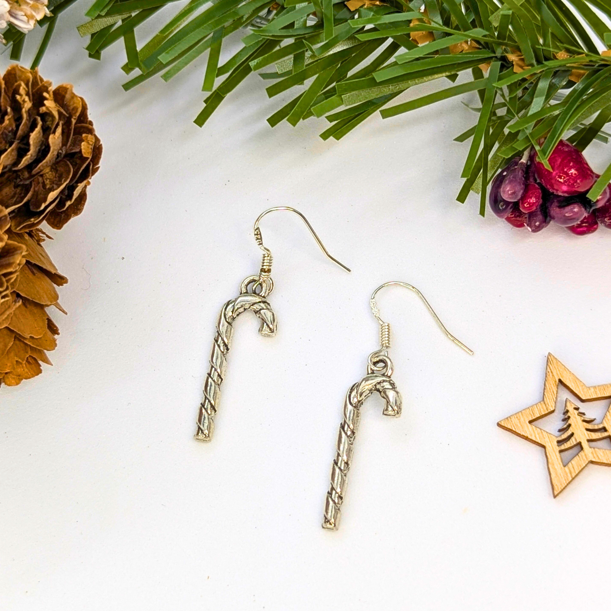 The Classic Candy Cane Earrings by Silver Sunbird rest on a white surface amid pine needles, a pine cone, red berries, and a wooden star—festive Christmas jewelry for the holiday season.