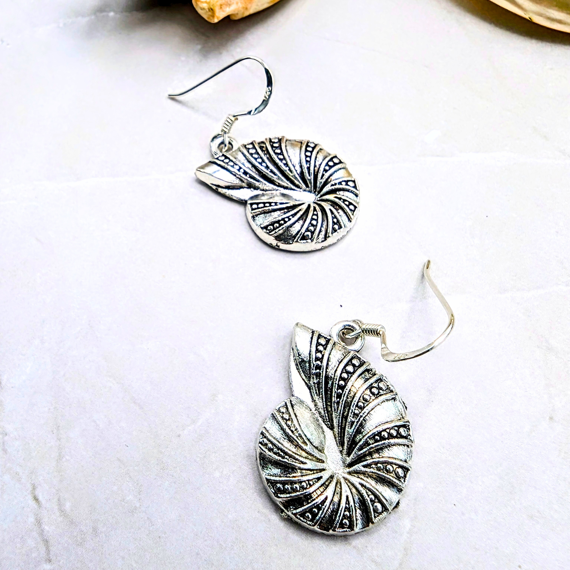 Shoreline Spiral Shell Earrings by Silver Sunbird—stylized nautilus designs with intricate textures, displayed on a light surface. Perfect ocean-inspired jewelry to elevate any look.