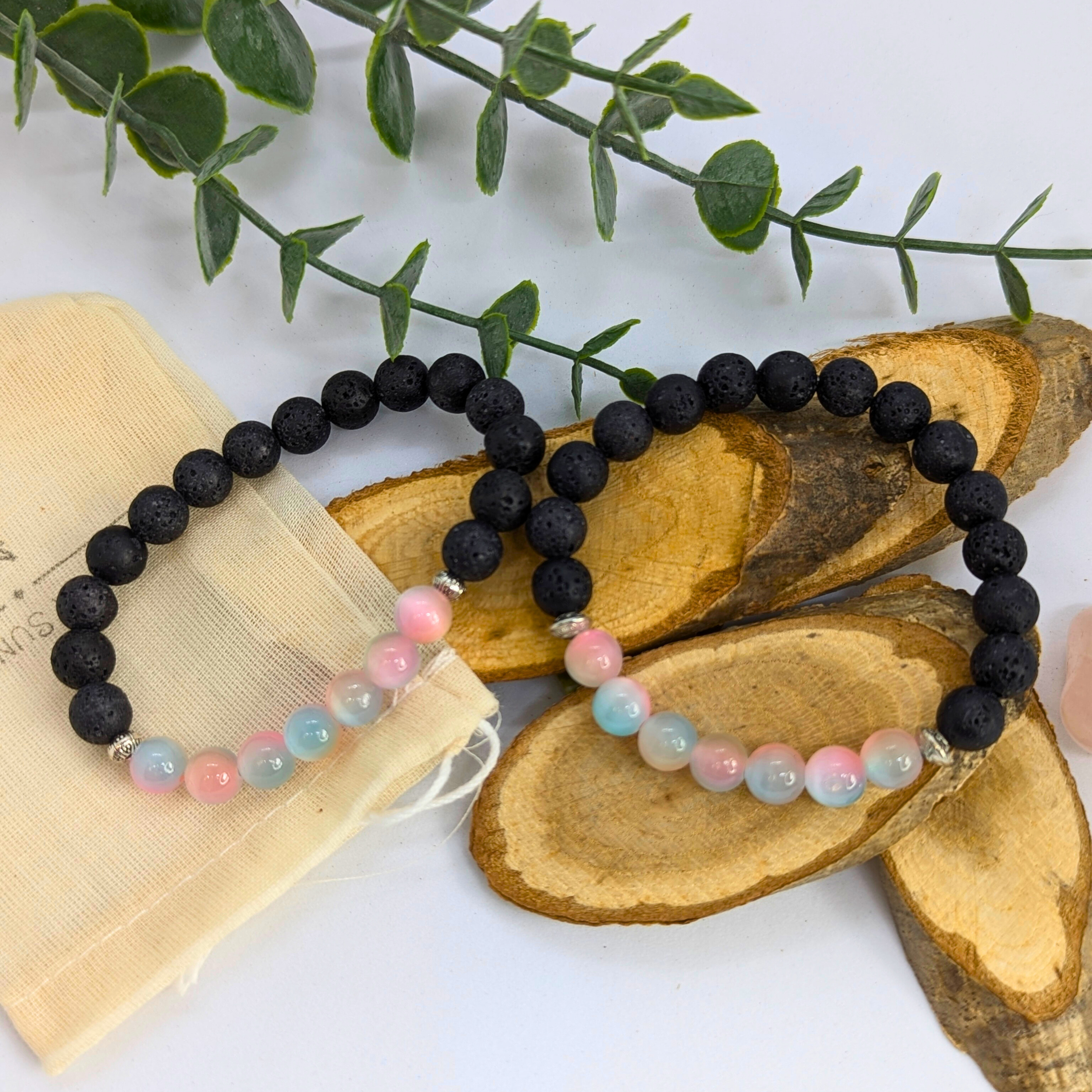 The Silver Sunbird Mermaid-Inspired Lava Stone Bracelet and a pastel beaded bracelet are displayed on wooden slices with green leaves and a beige fabric pouch in the background—an ideal set for ocean lovers.
