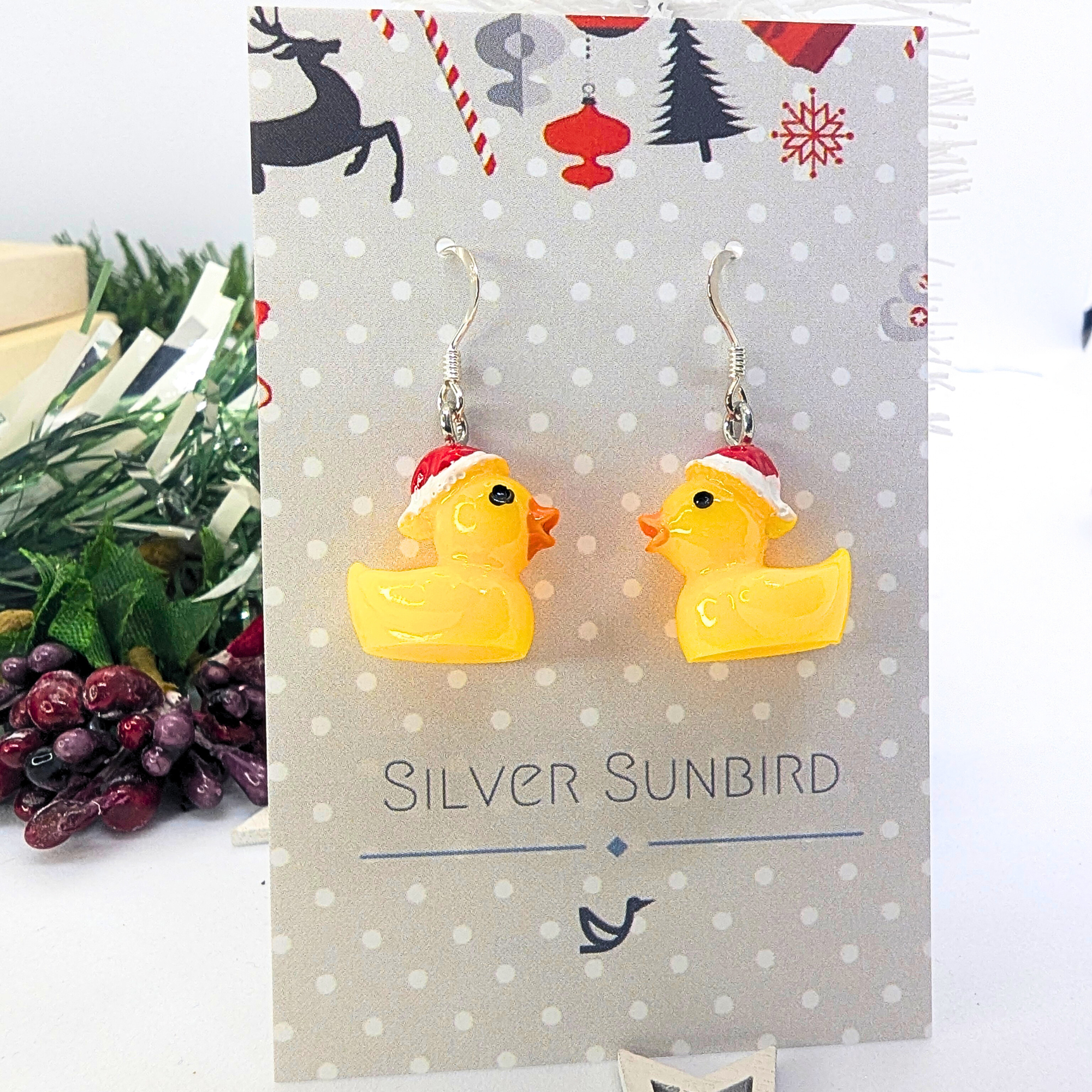 The Festive Duck Earrings from Silver Sunbird are yellow ducks adorned with red and white Santa hats, showcased on a card featuring the brand&