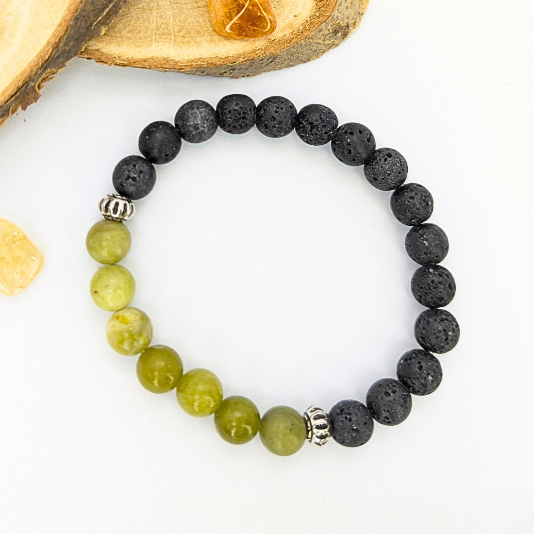 Silver Sunbird’s Peridot and Lava Stone Bracelets feature alternating green peridot and black lava beads, accented with two silver spacers. Arranged in a circle on a white surface near wood pieces and yellow stones.
