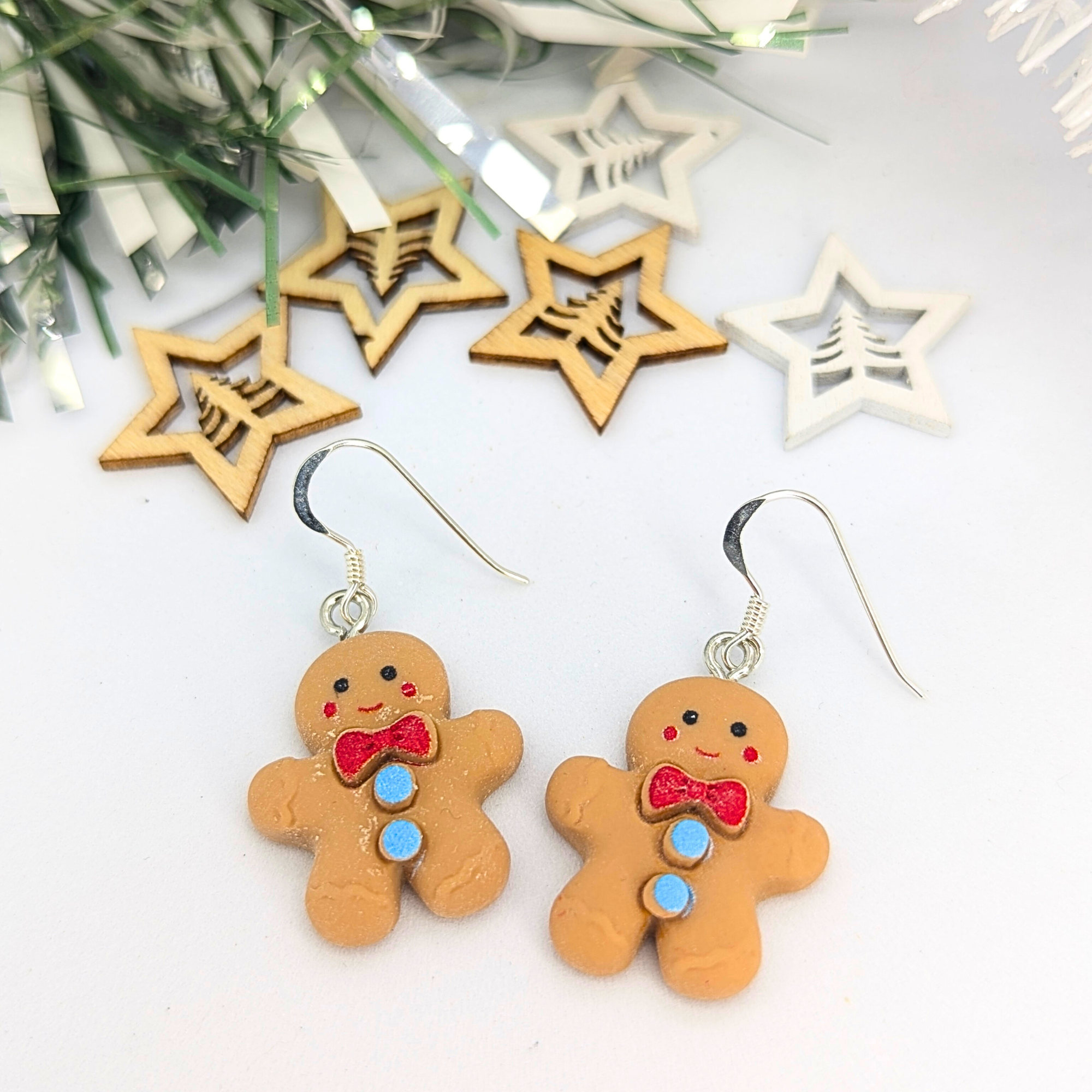 Cheerful Gingerbread Man Earrings by Silver Sunbird, featuring red bows and blue buttons, are showcased amidst wooden star ornaments and festive decor.