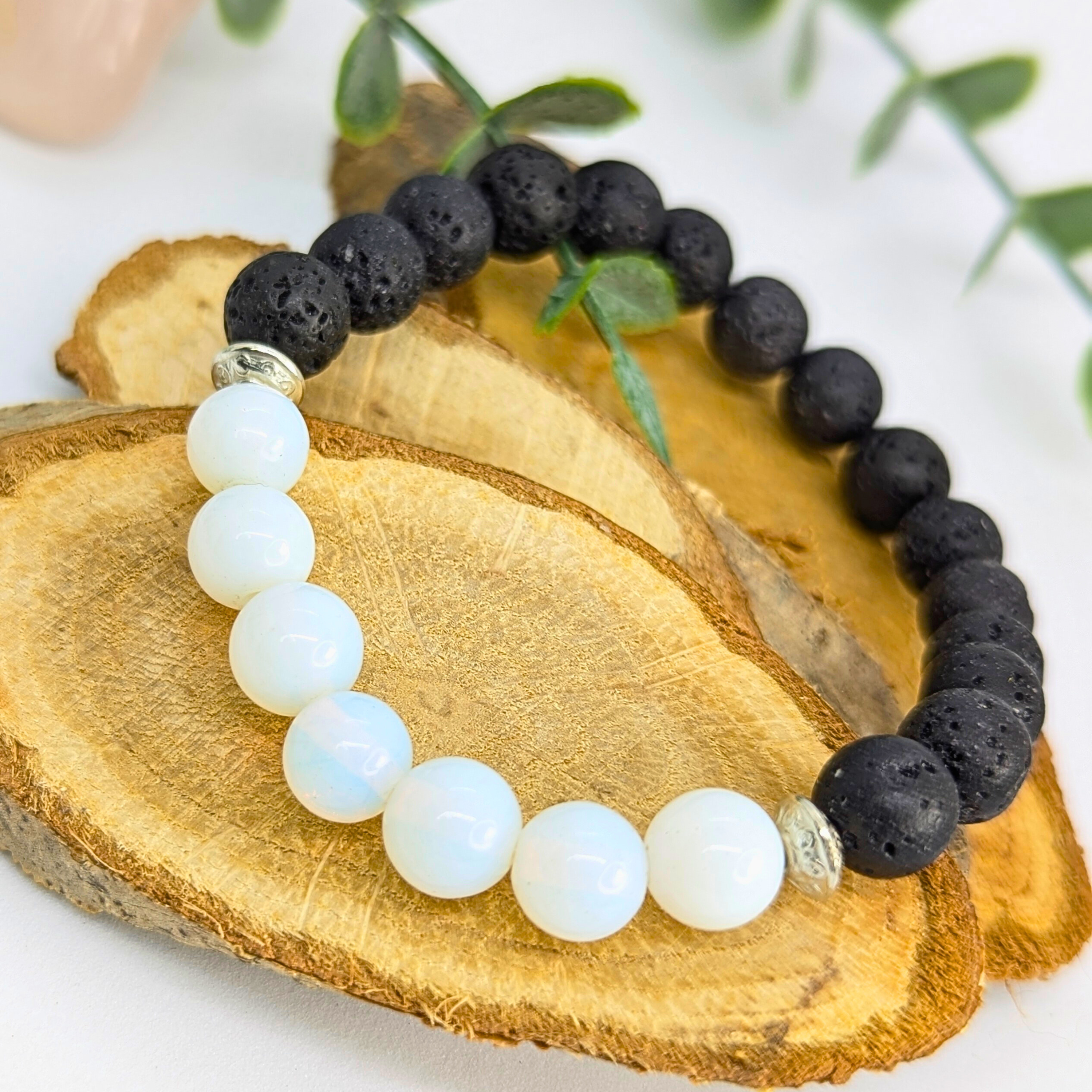 The Silver Sunbird Opal and Lava Stone Bracelets feature alternating glossy white opal and matte black lava beads, shown on a wooden surface with green leaves in the background.