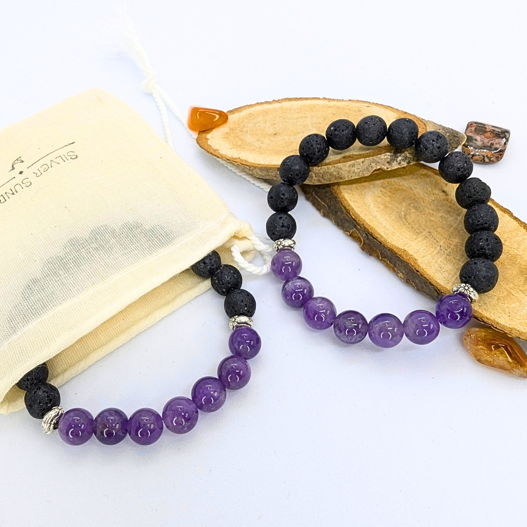 Two stretch bracelets featuring black lava stones and purple amethyst beads are displayed on a wooden slice. A beige drawstring pouch with "Silver Surfer" branding is positioned to the side, along with small polished stones in orange and brown hues. The amethyst beads are glossy and smooth, contrasting with the porous texture of the lava stones.