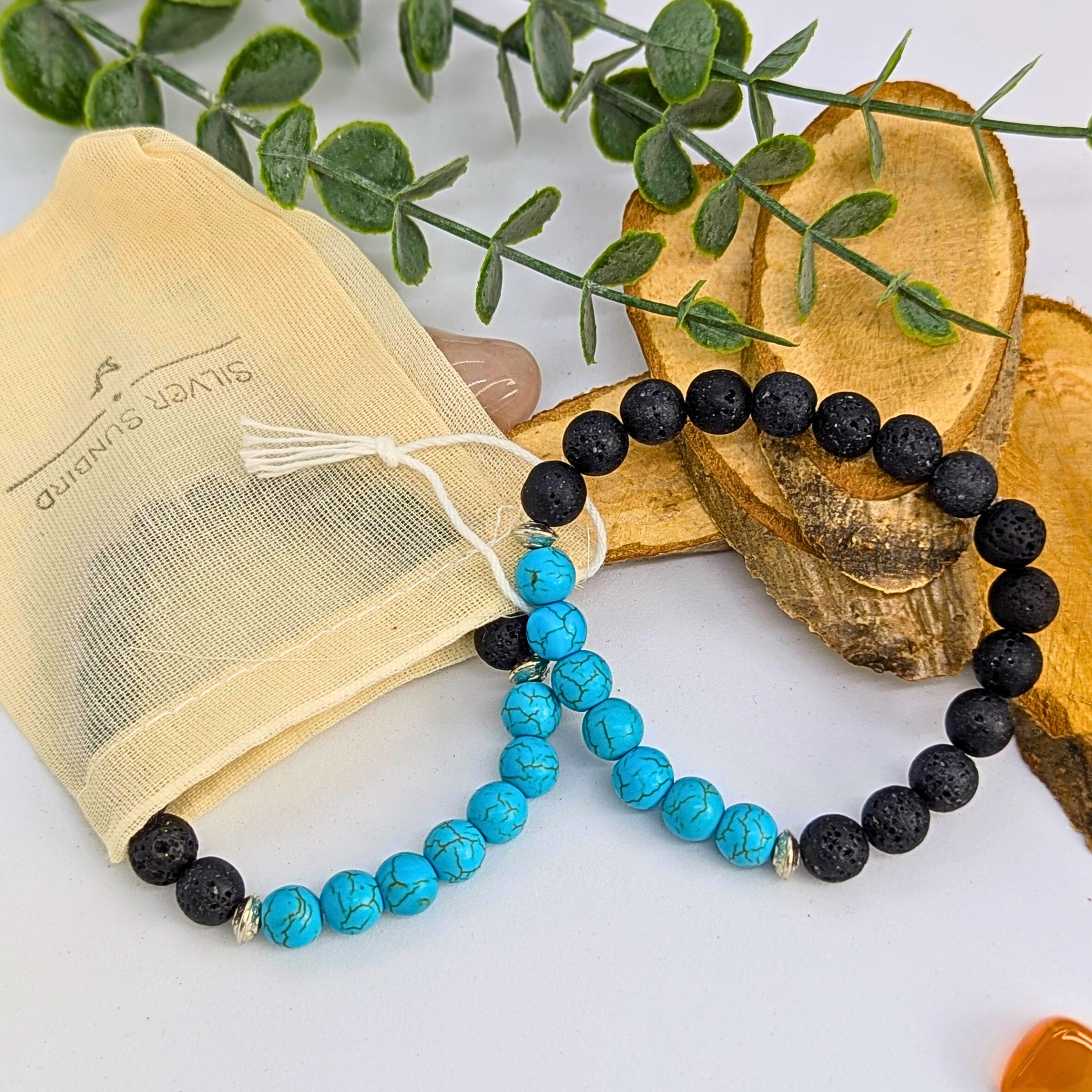 Silver Sunbird’s Turquoise and Lava Stone Bracelets are shown beside a beige drawstring pouch, wooden slices, and green leaves on a white surface.