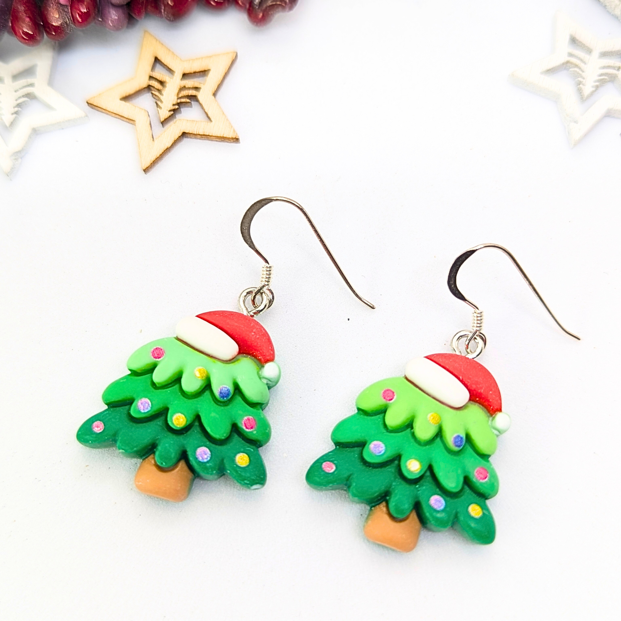 Christmas Tree Earrings by Silver Sunbird feature a festive design with colorful decorations and Santa hat toppers. Displayed against a white background, these holiday accessories are beautifully complemented by nearby wooden star decorations.