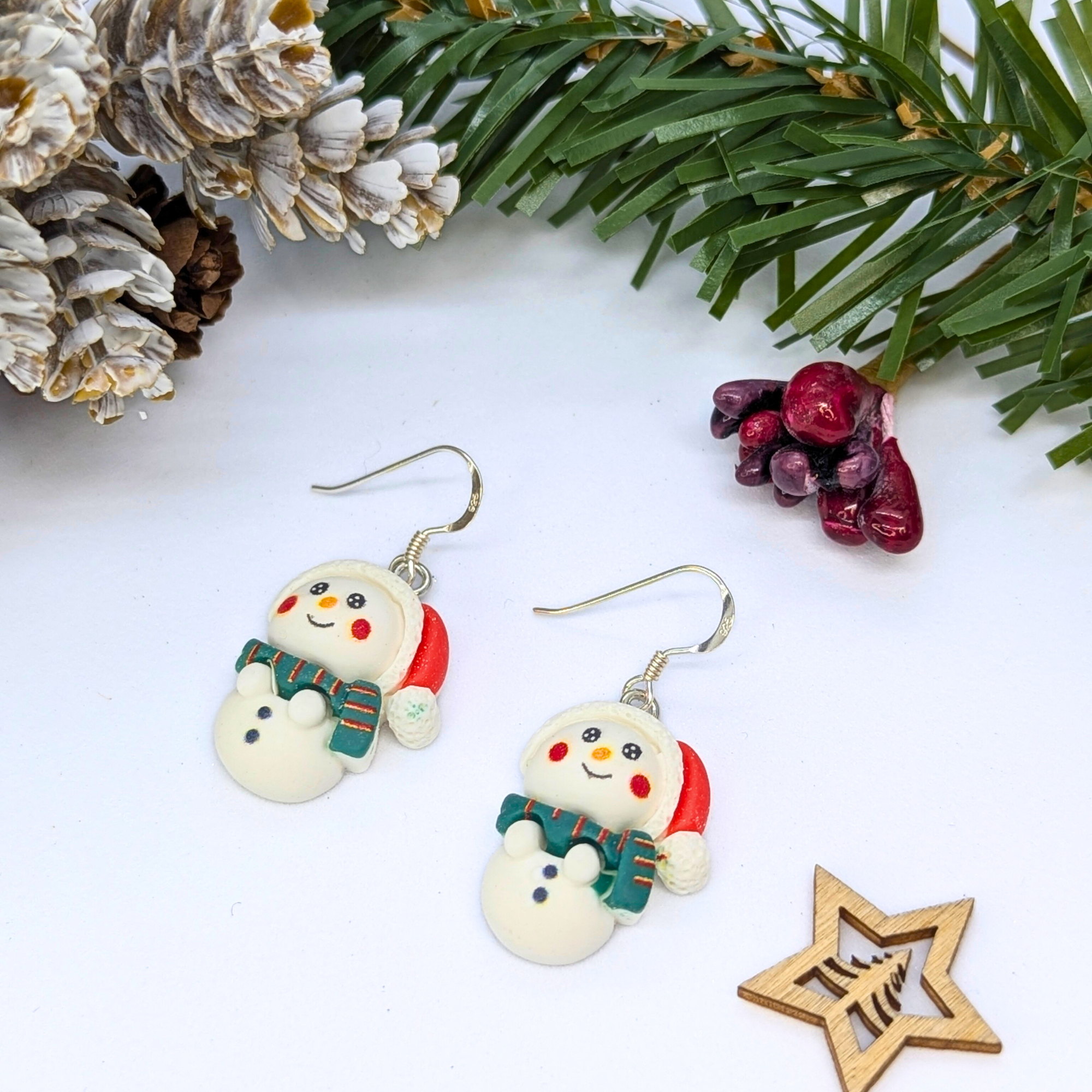 Bright Snowmen Earrings by Silver Sunbird feature charming snowman shapes with red hats and green scarves, displayed on a white surface beside pine branches and festive decor—the perfect holiday accessory for your Christmas jewelry collection.