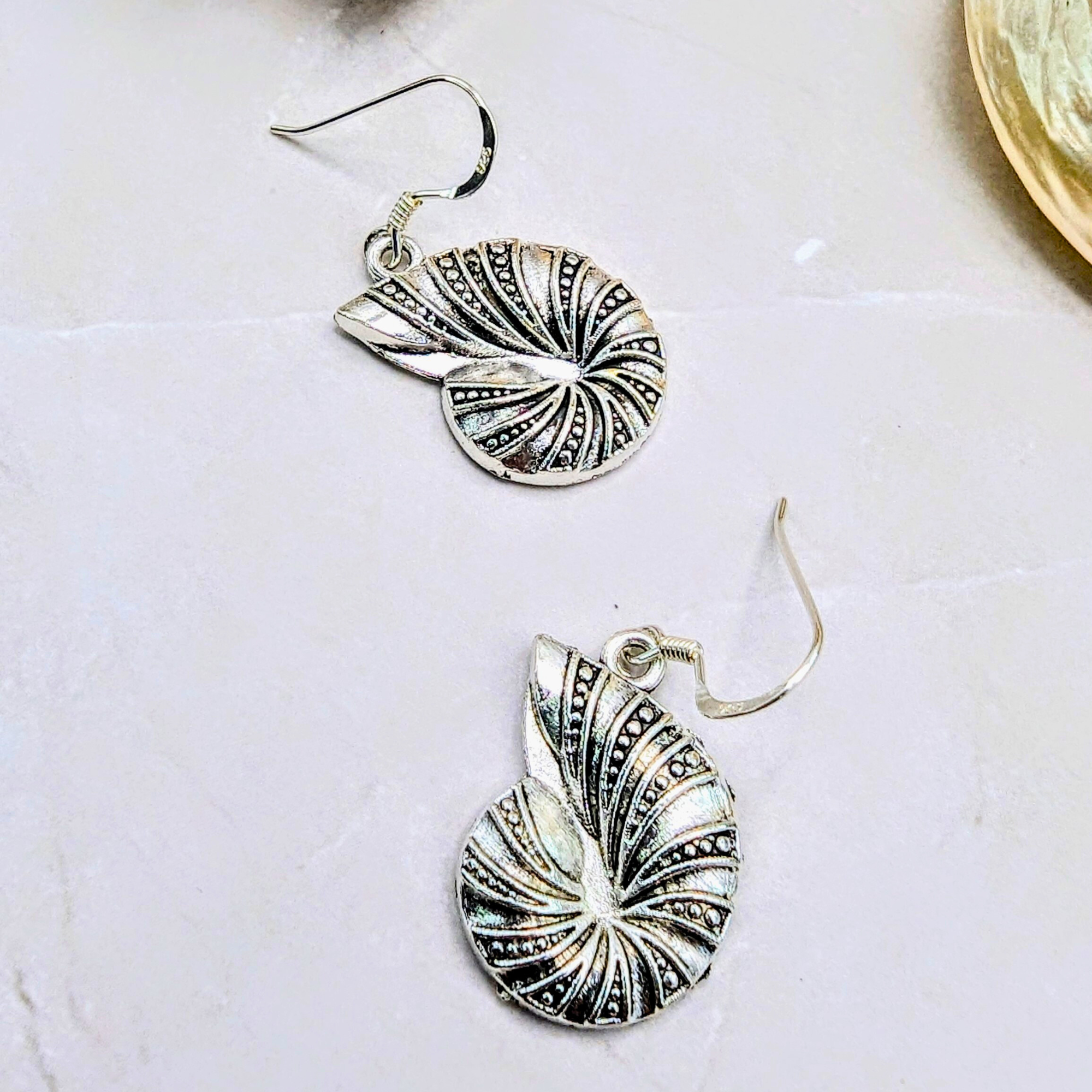 The Silver Sunbird Shoreline Spiral Shell Earrings, inspired by the ocean and shaped like nautilus shells with engraved lines and dot patterns, are displayed on a light-colored stone surface.