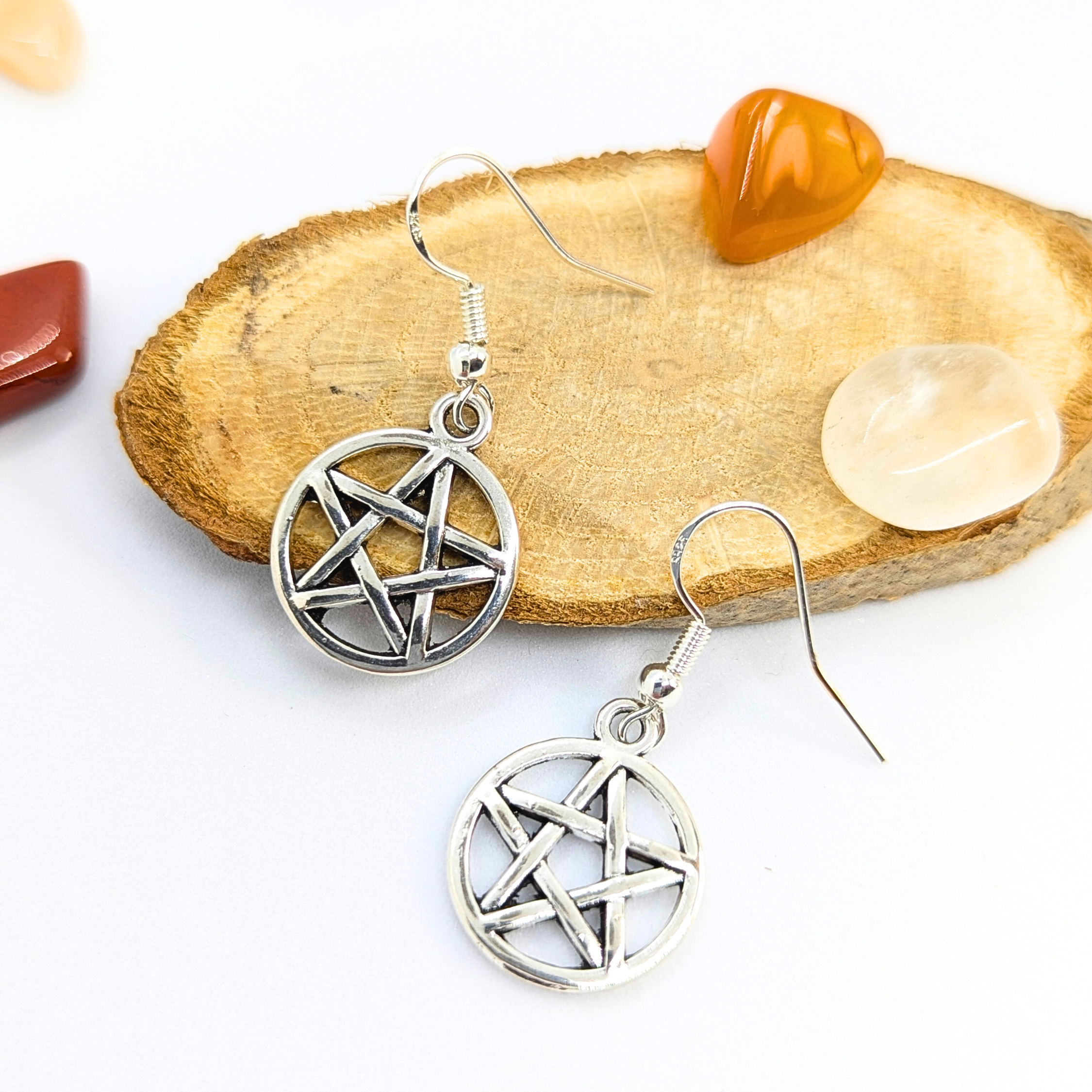Silver Sunbird Pentagram Earrings, ideal Wicca jewelry, shown on a wooden slice surrounded by polished red, orange, and clear stones against a white background.