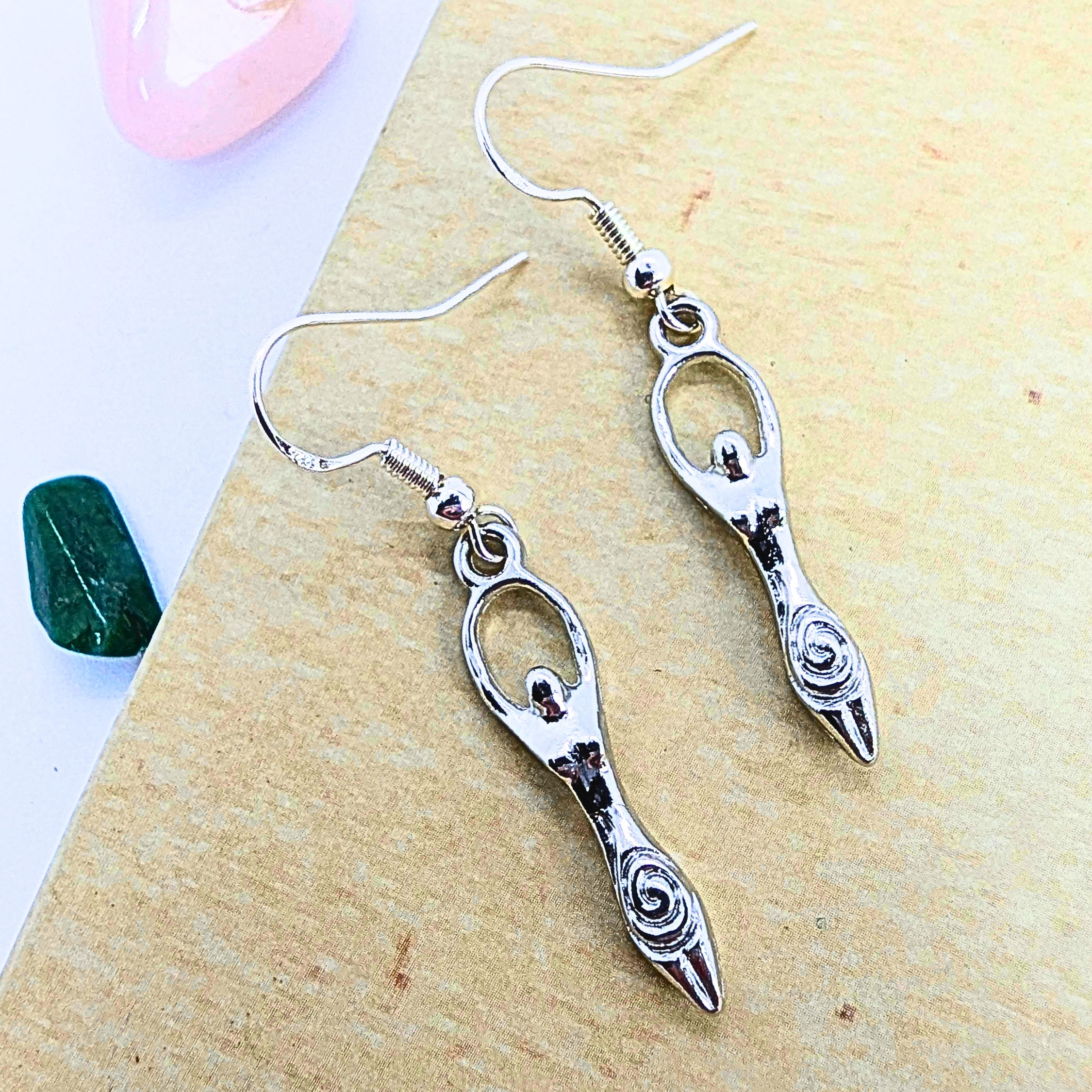 Silver Sunbird’s Fertility Goddess Dangle Earrings in silver feature spiral designs at the base, showcased on a tan surface with a pink stone and a green stone in the background.