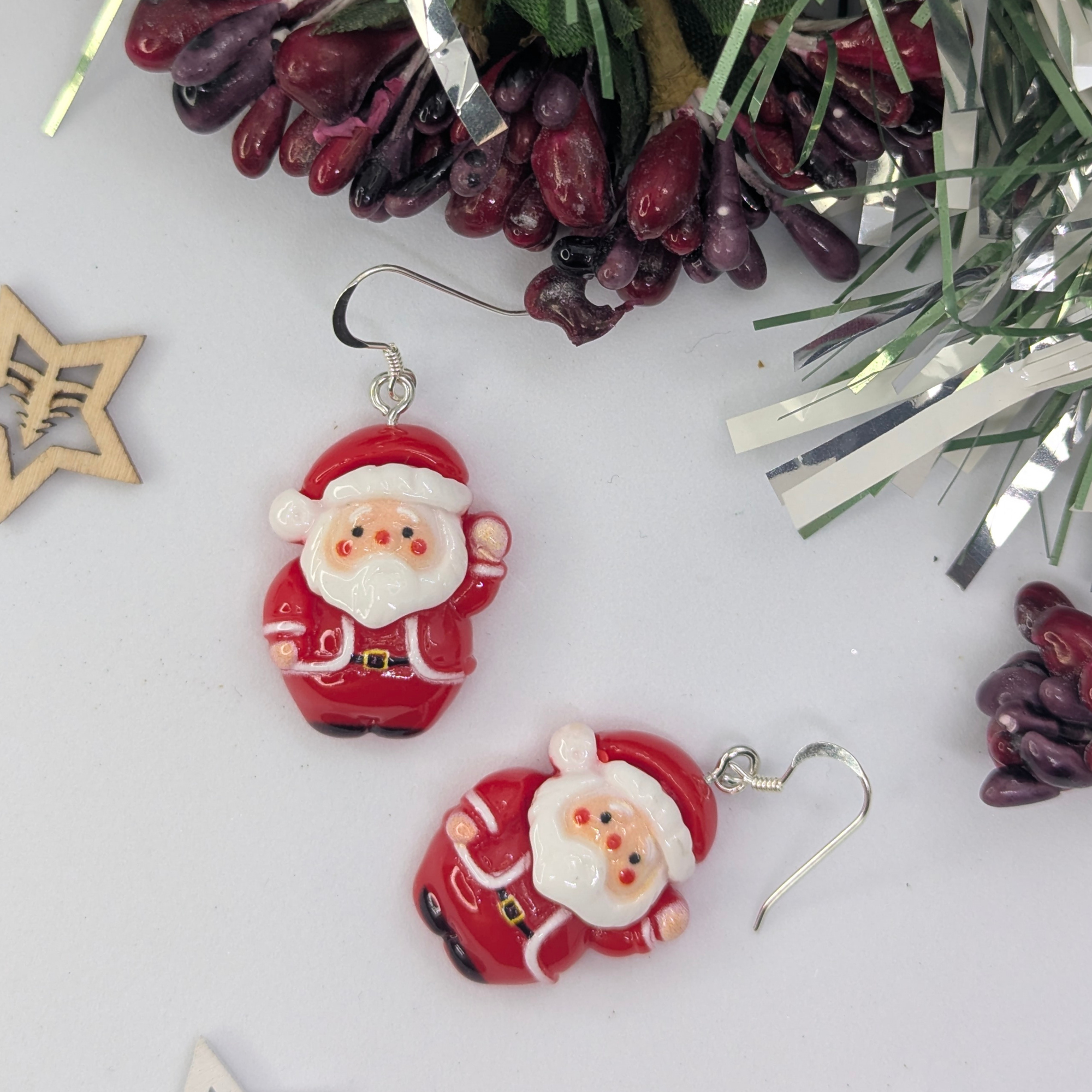 The Jolly Santa Earrings by Silver Sunbird rest on a white surface, surrounded by festive decorations like berries and shimmering silver and gold tinsel. These charming Christmas accessories add a playful touch to your festive jewelry collection.