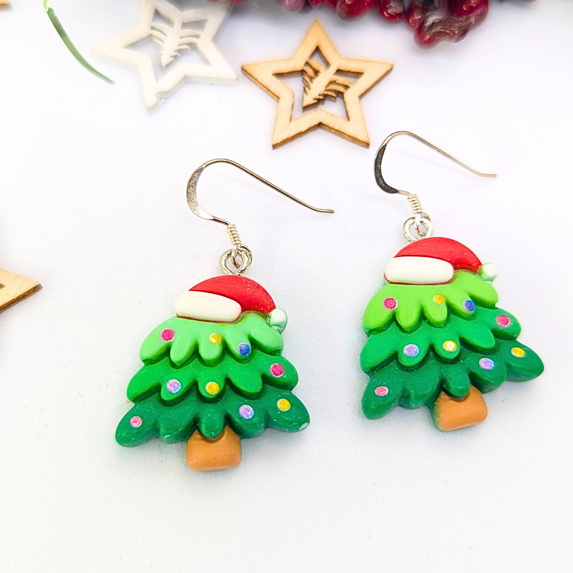 The Christmas Tree Earrings by Silver Sunbird are the perfect holiday accessory, featuring festive adornments with Santa hats and colorful ornaments. Exhibited on a white surface, they shimmer brilliantly beside wooden star decorations, embodying the spirit of seasonal jewelry.