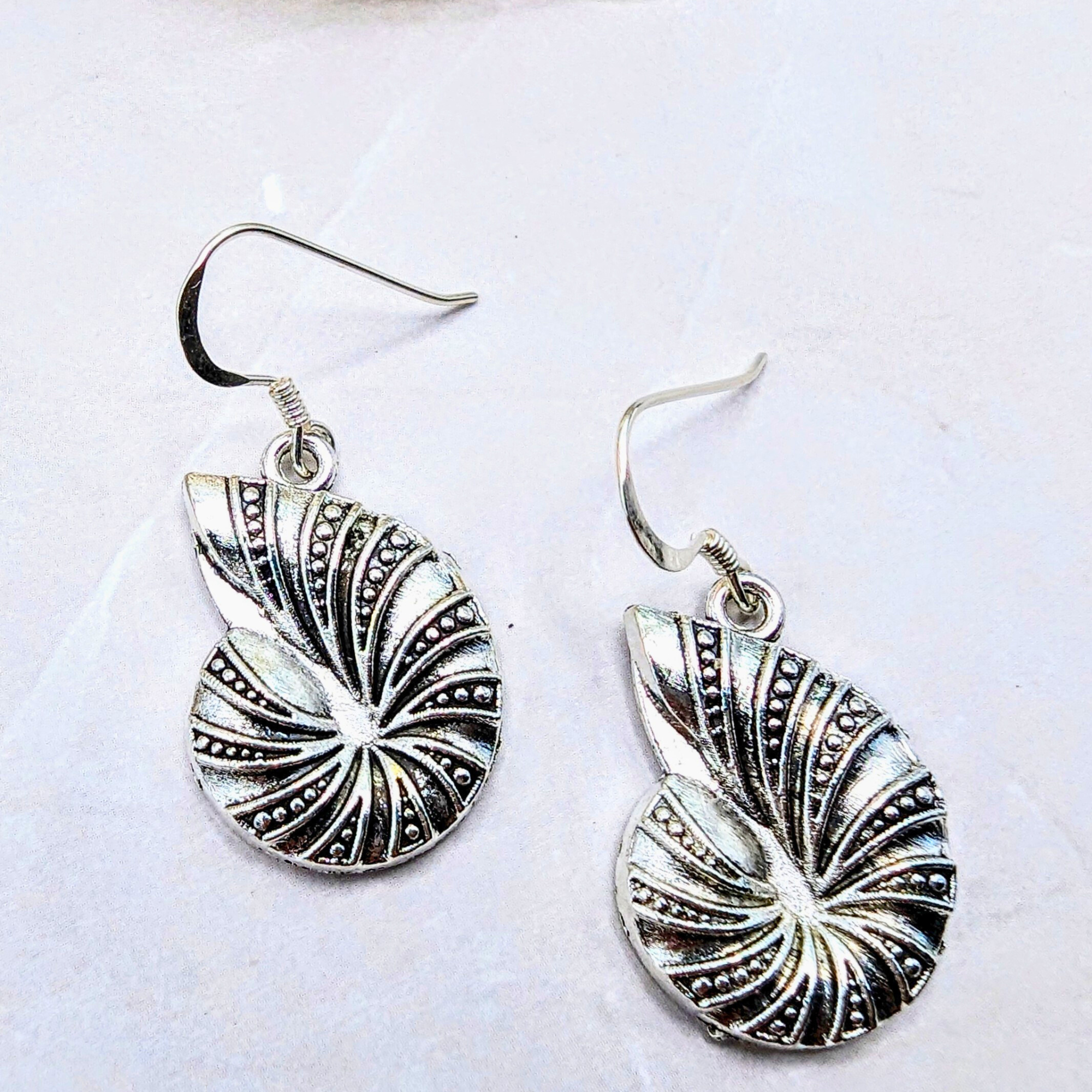 The Silver Sunbird Shoreline Spiral Shell Earrings feature stylized sterling silver seashells with embossed patterns and dot details—ideal for ocean-inspired jewellery lovers. Displayed on a white background.