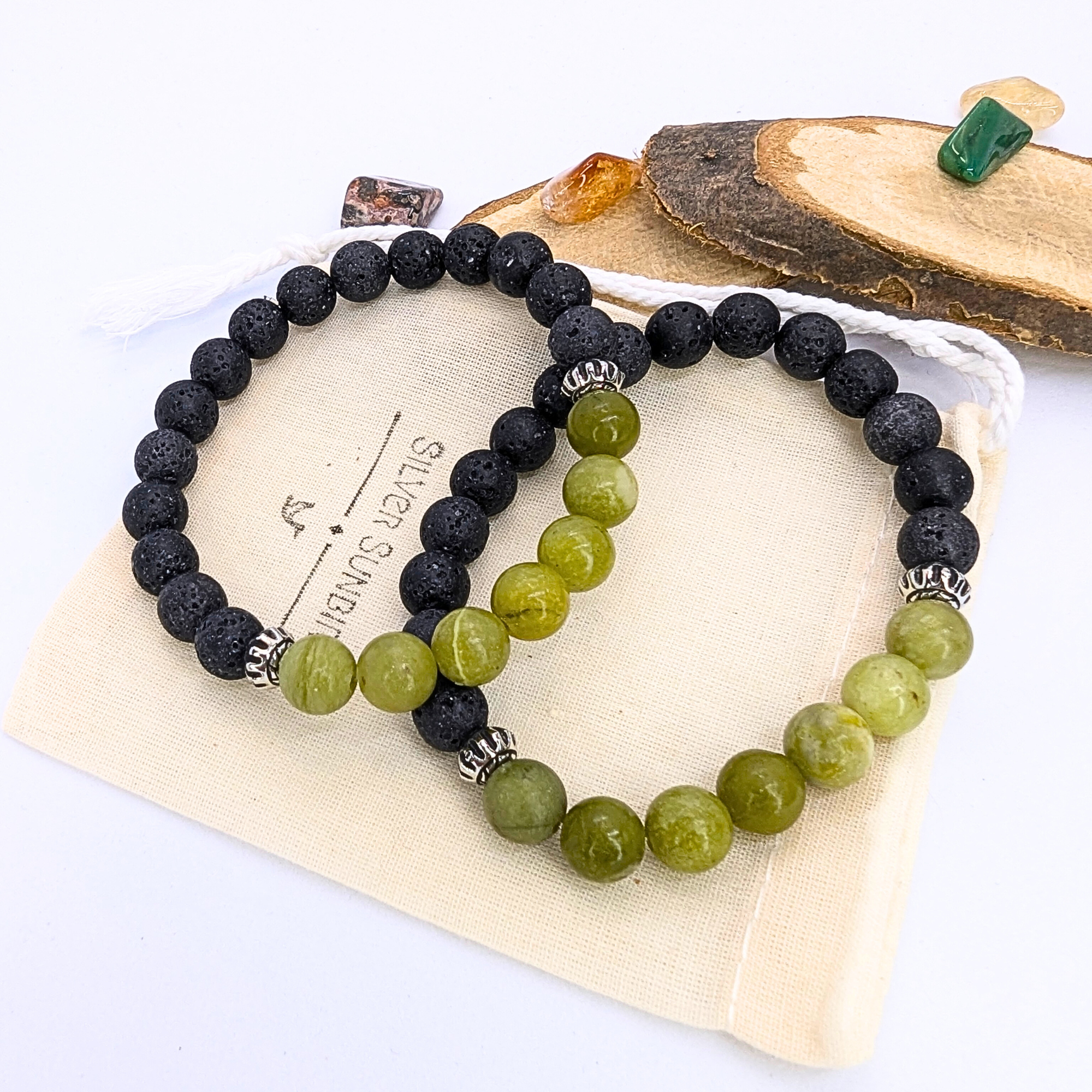 The Peridot and Lava Stone Bracelets by Silver Sunbird—one with black lava stones, the other with green peridot and silver accents—are displayed on a beige pouch, accented by decorative stones and a wooden slab.