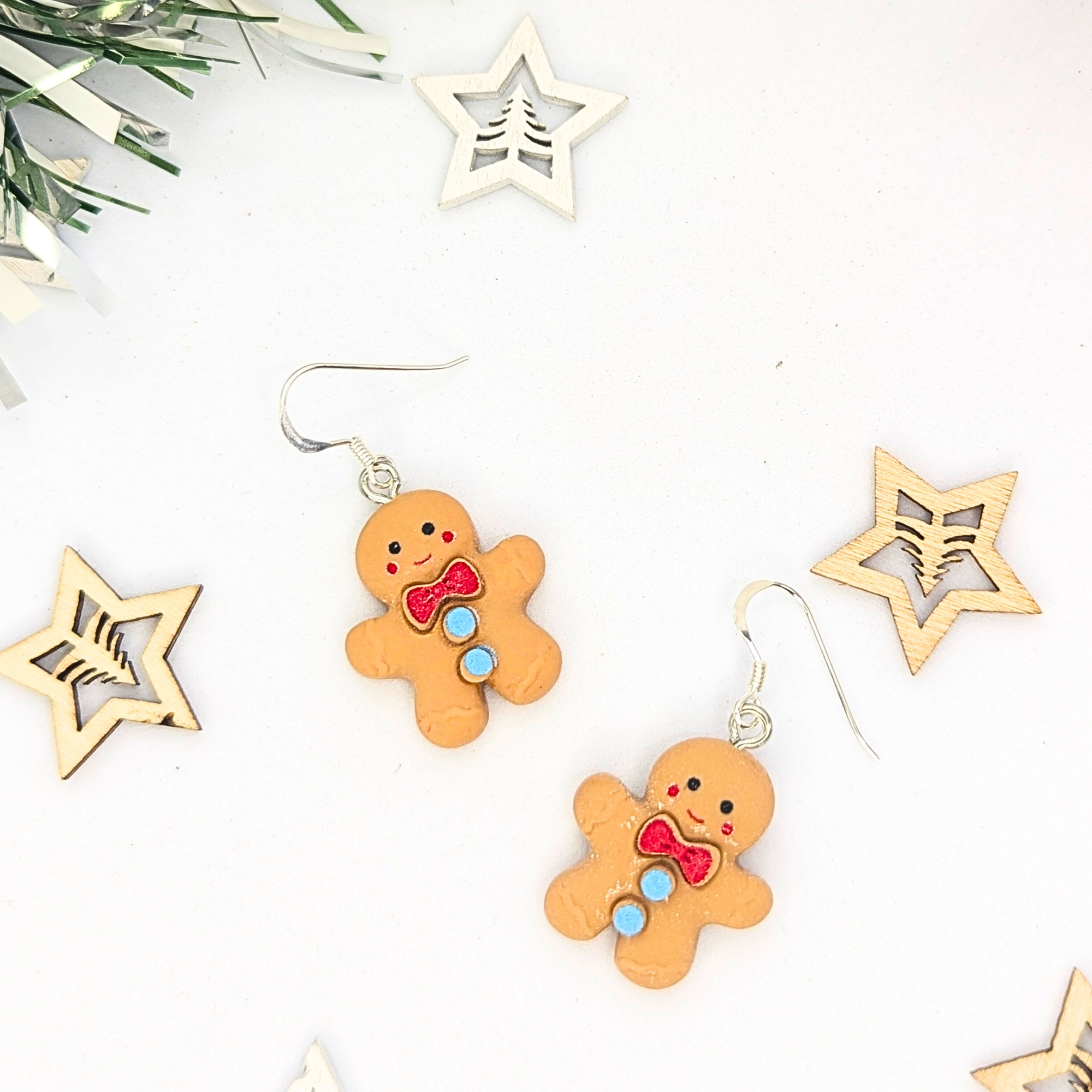 The Cheerful Gingerbread Man Earrings by Silver Sunbird feature gingerbread men adorned with red bows and colorful buttons against a white background. They are complemented by decorative wooden stars and greenery, enhancing the holiday theme.