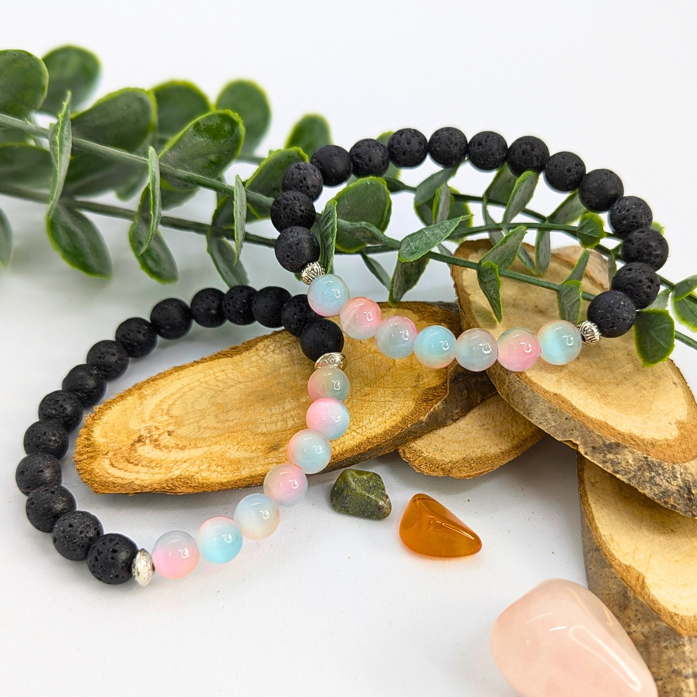 Two beaded bracelets rest on wooden slices with green leaves. One is the Silver Sunbird Mermaid-Inspired Lava Stone Bracelet; the other is a pastel bracelet in black, pink, and blue. Nearby lie small polished stones in green, orange, and pink.