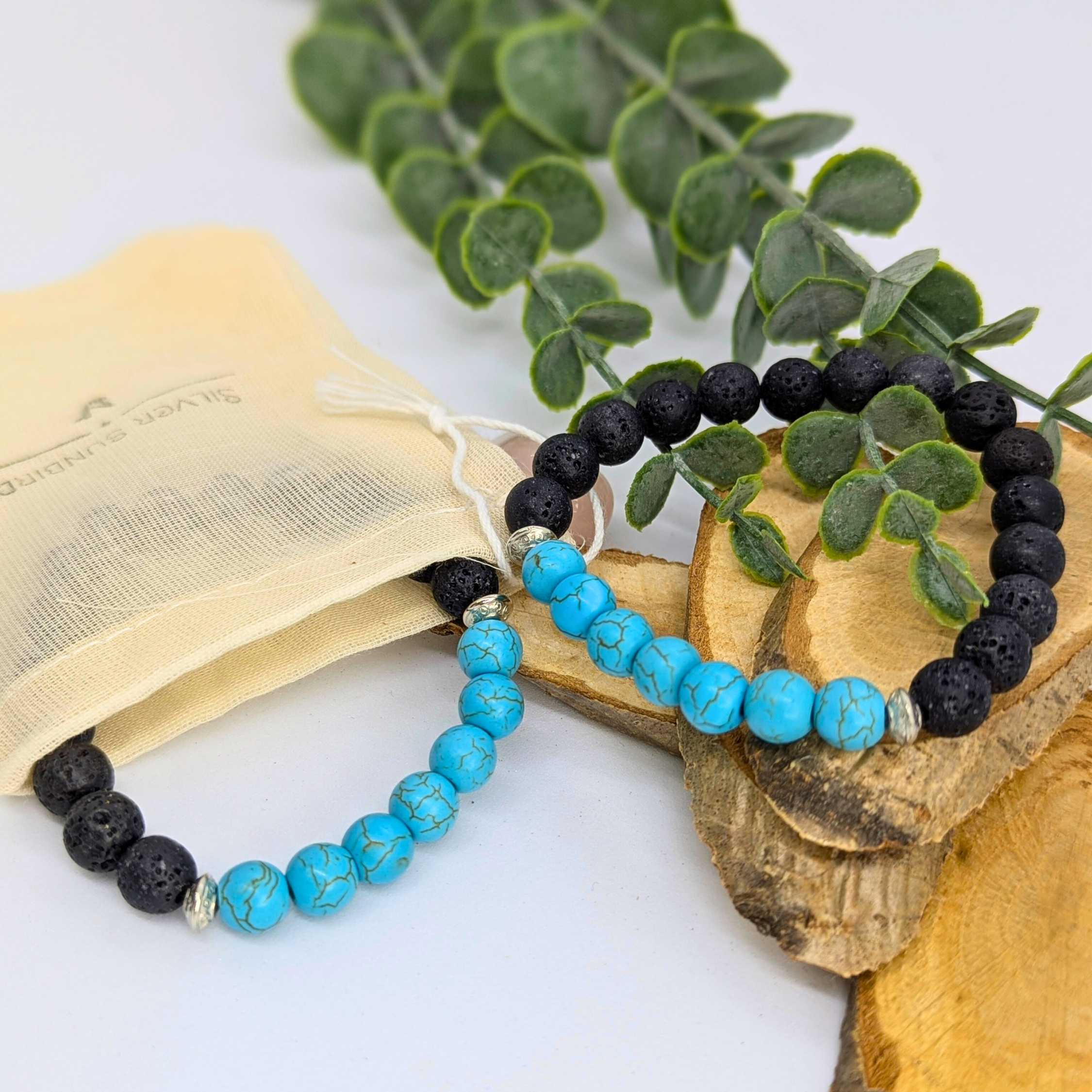 The Silver Sunbird Turquoise and Lava Stone Bracelets, featuring black, blue, and turquoise beads, are displayed on wooden slices beside greenery and a beige drawstring pouch.