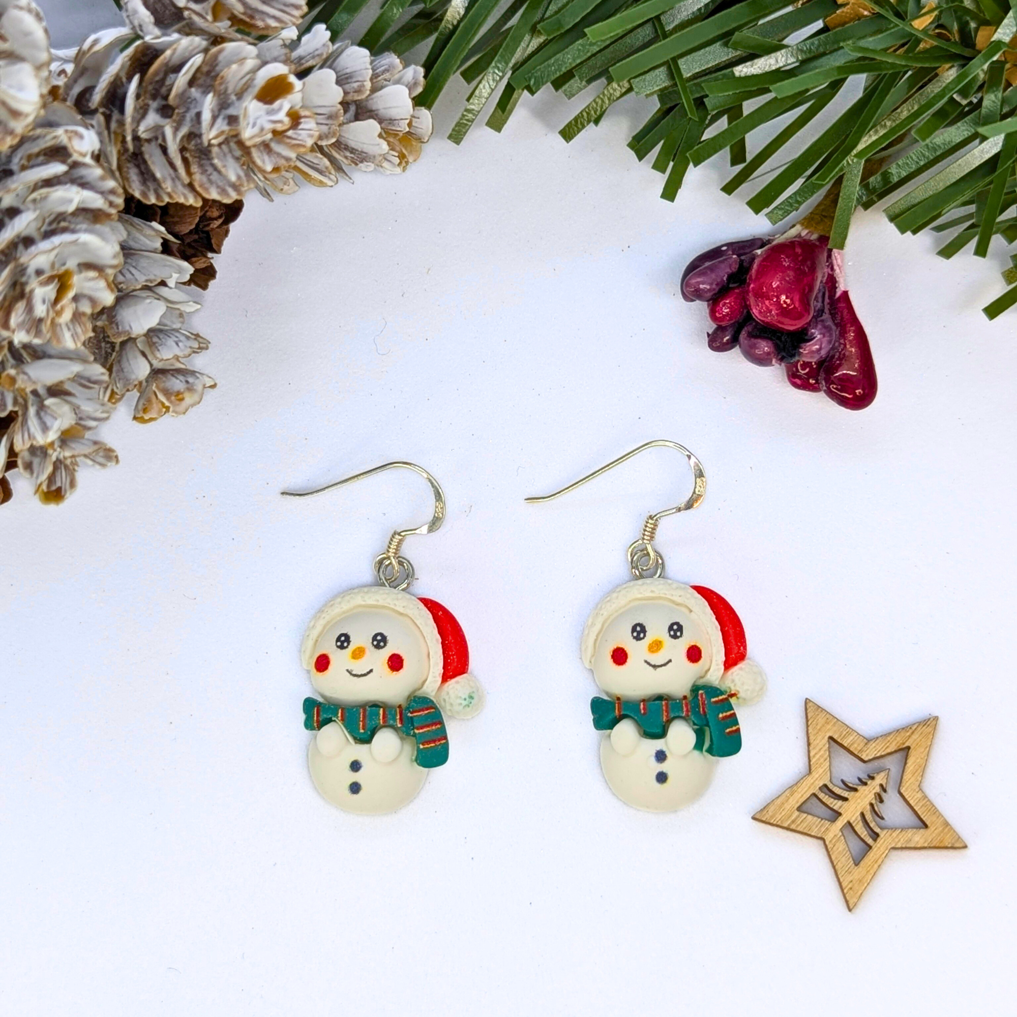 Bright Snowmen Earrings by Silver Sunbird feature festive snowmen with Santa hats and green scarves, displayed on a white background amid pinecones, fir branches, red berries, and a wooden star—perfect Christmas jewelry.