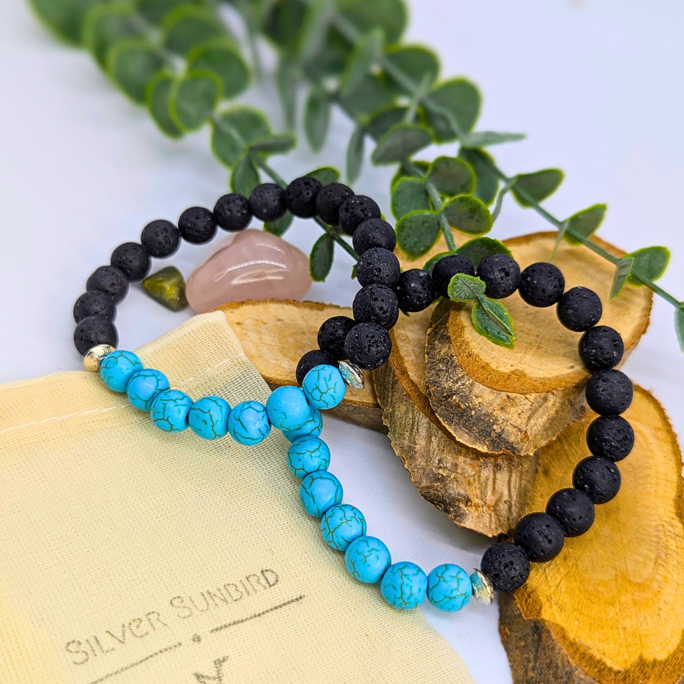 Silver Sunbird’s Turquoise and Lava Stone Bracelets are displayed in heart shapes on wooden slices, with greenery, a rose quartz stone, and a beige pouch nearby.