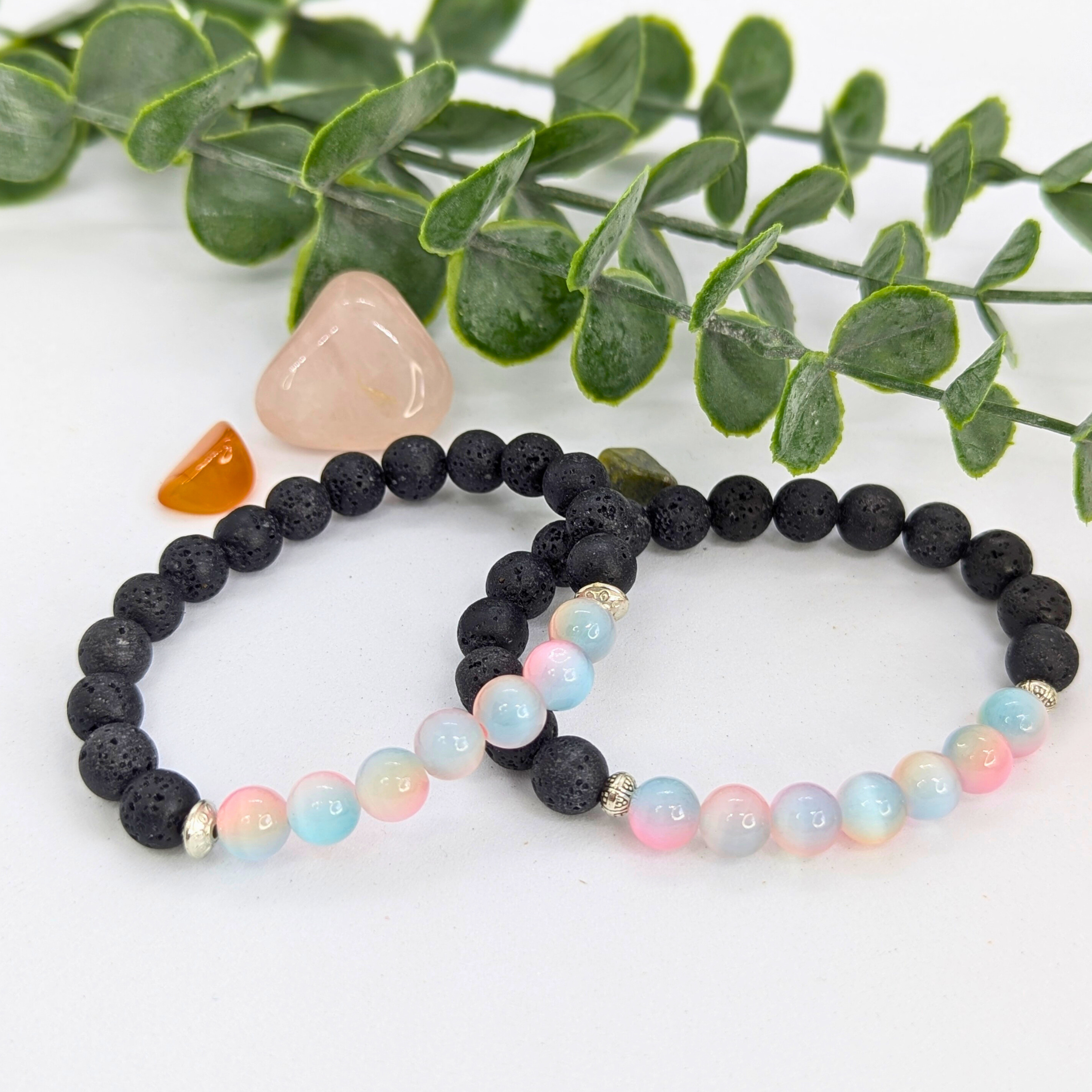 Two beaded bracelets rest on a white surface: the Silver Sunbird Mermaid-Inspired Lava Stone Bracelet with black lava beads and a pastel bracelet of black, blue, and pink. Green leaves and stones accent the scene—perfect for ocean lovers.