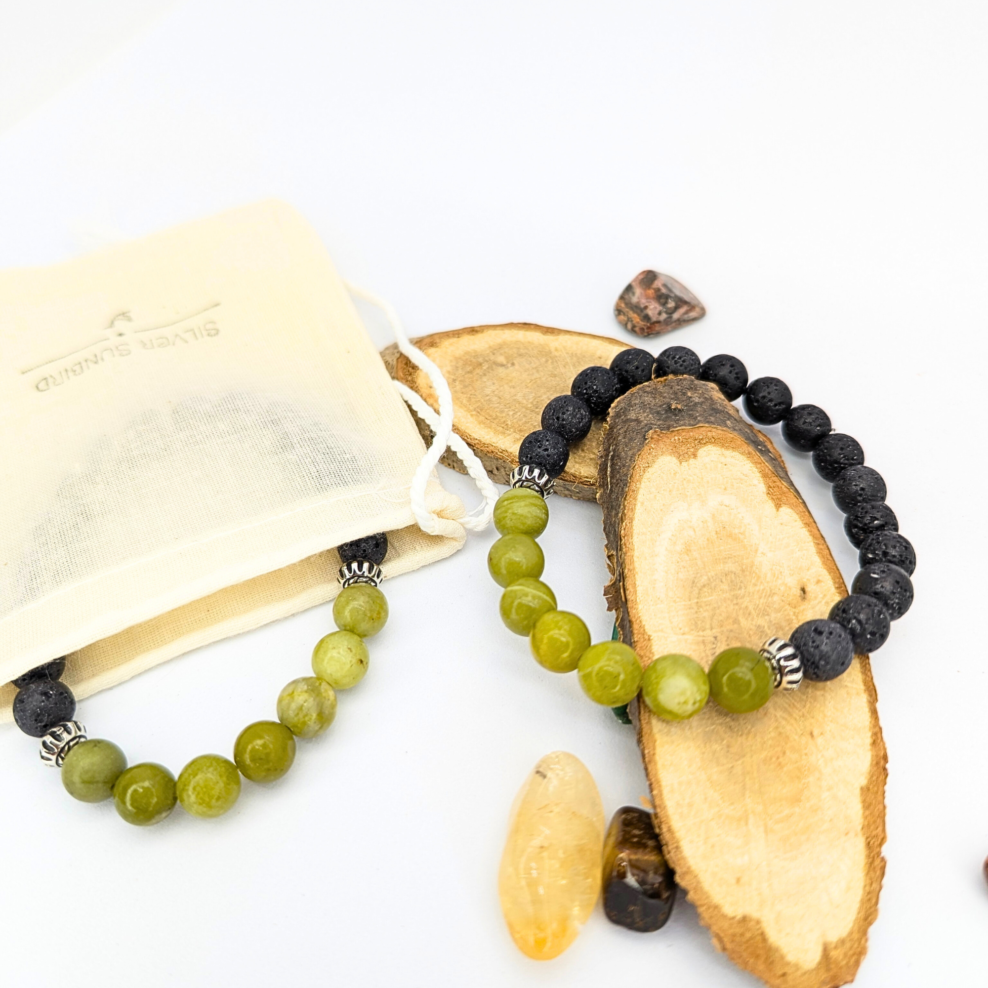 Two Peridot and Lava Stone Bracelets by Silver Sunbird—one with black lava beads and silver accents, the other with green peridot beads—rest on wood slices, alongside crystals and a cream drawstring pouch on a white background.