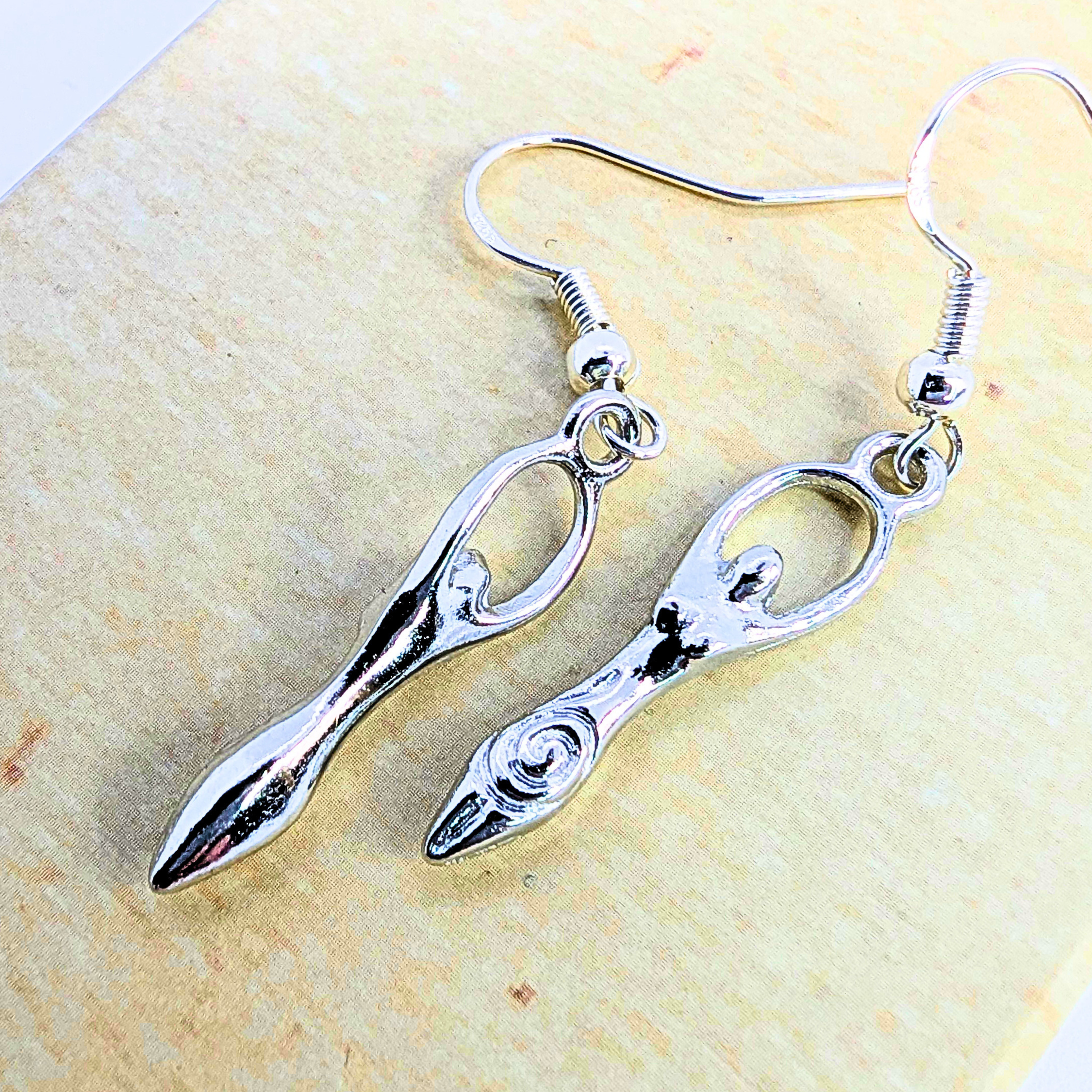 Fertility Goddess Dangle Earrings by Silver Sunbird—sterling silver, stylized goddess figures with arms raised, displayed on a light beige surface. Unique symbolic jewelry to enhance any collection.
