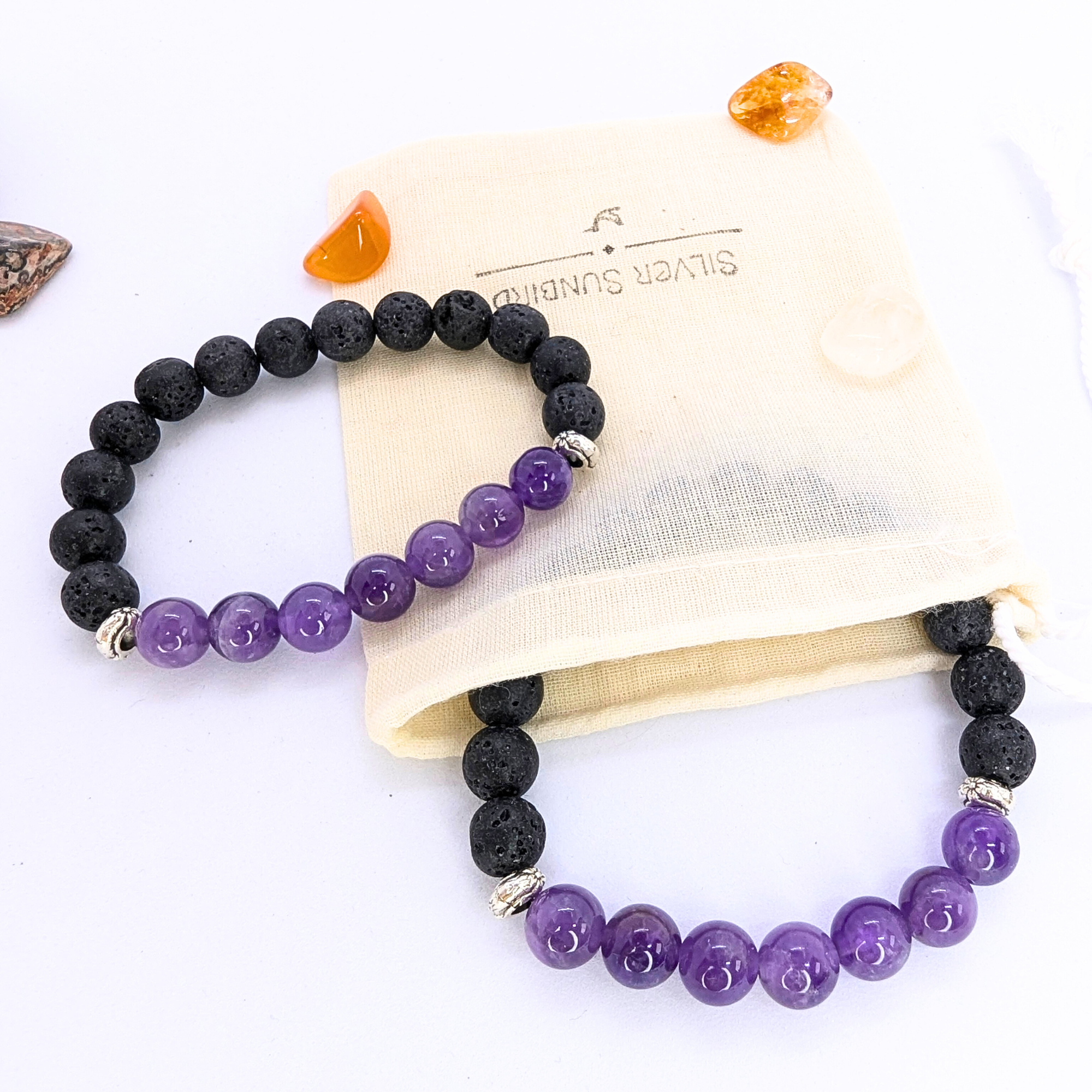 Two stretch bracelets featuring black lava stones and purple amethyst beads are displayed on a wooden slice. A beige drawstring pouch with "Silver Surfer" branding is positioned to the side, along with small polished stones in orange and brown hues. The amethyst beads are glossy and smooth, contrasting with the porous texture of the lava stones.