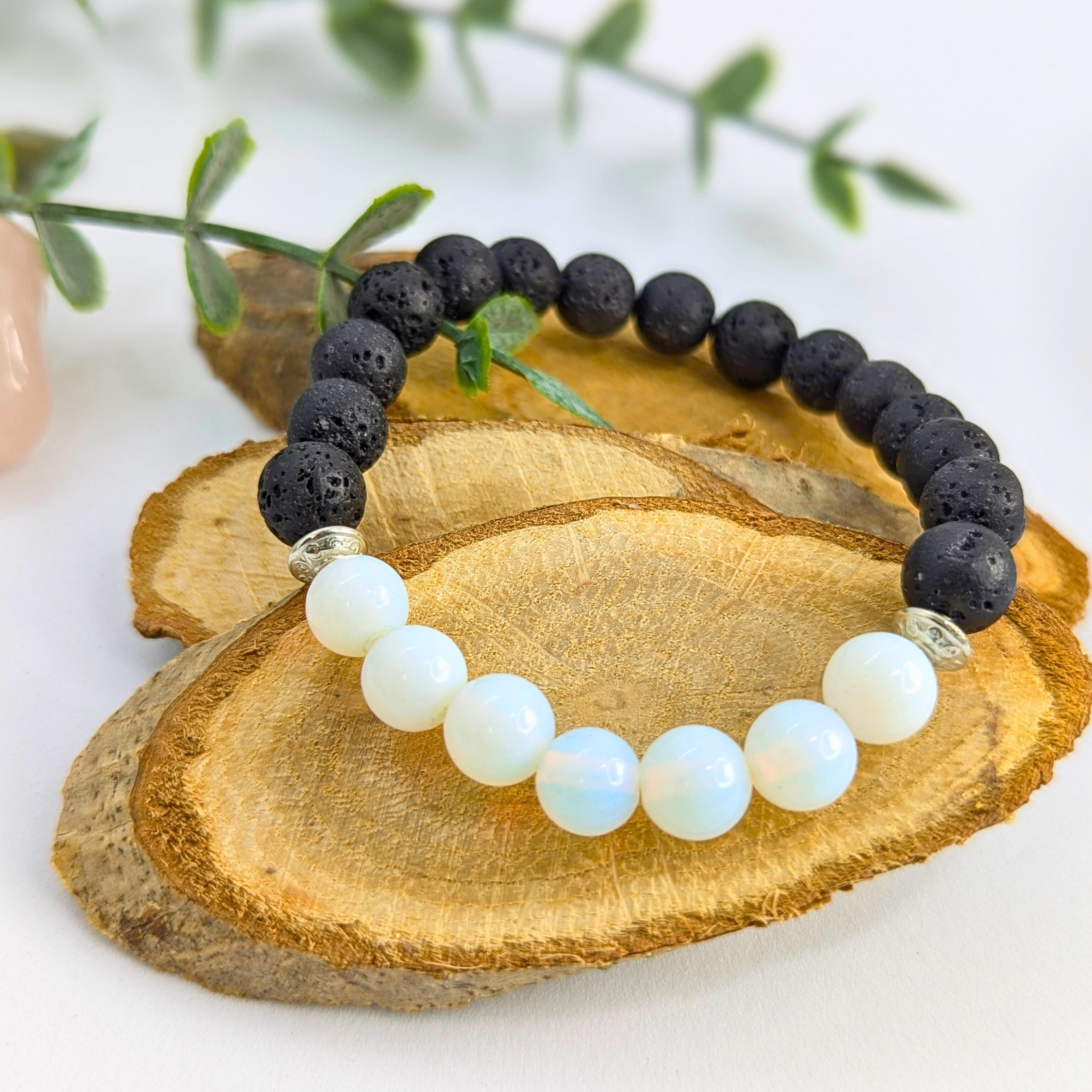 The Silver Sunbird Opal and Lava Stone Bracelets, featuring black lava stones and white opalite beads, are displayed on a wooden slab with green leaves in the background.