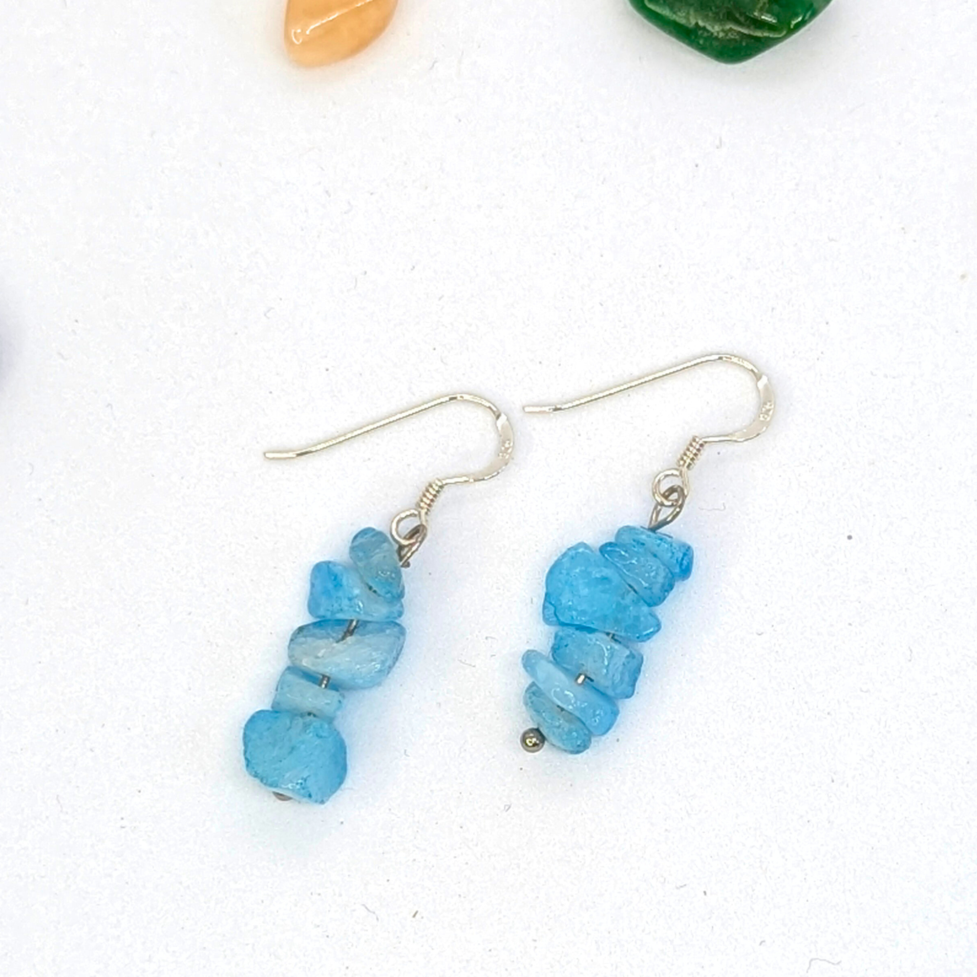 Silver Sunbird Aquamarine Gemstone Earrings feature handmade, stacked irregular blue aquamarine beads on wire hooks, shown on a white background with small orange and green stones beside them.