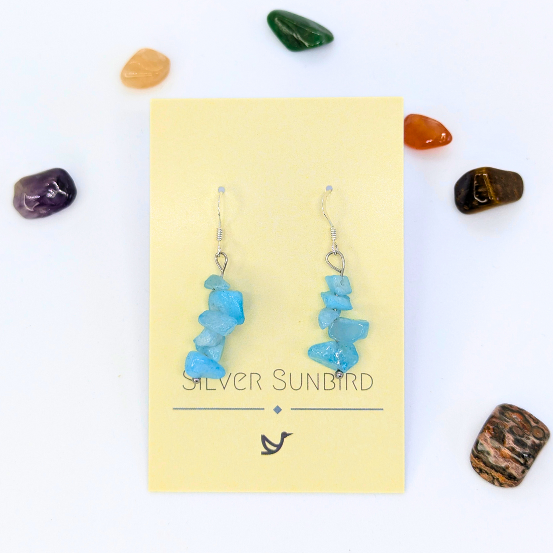 Aquamarine Gemstone Earrings by Silver Sunbird displayed on a yellow brand card, surrounded by colorful polished stones on a white background.