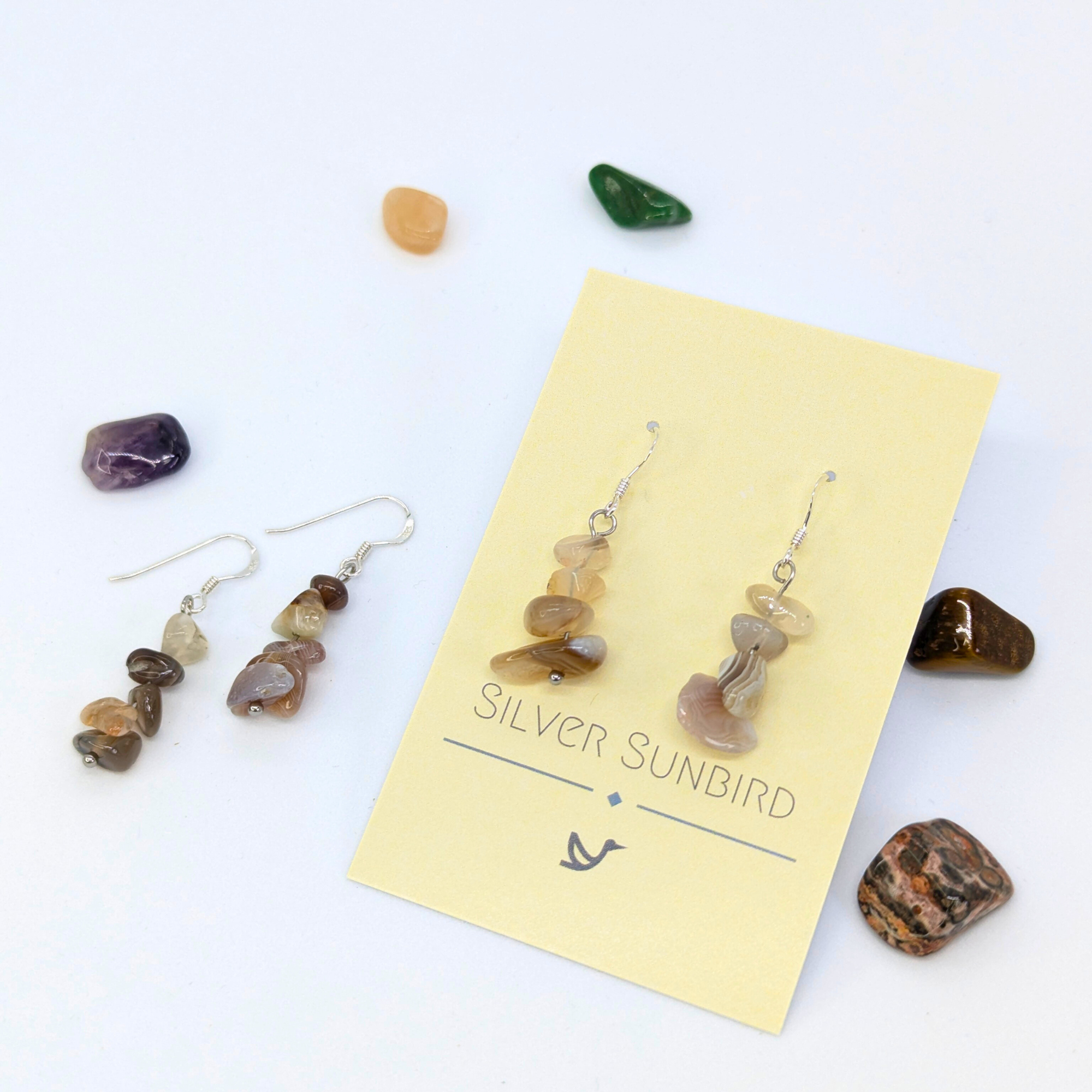 A pair of Silver Sunbird Botswana Agate Gemstone Earrings on a pale brand card, surrounded by colorful tumbled stones, with another set of sterling silver earrings beside the card on a white background.