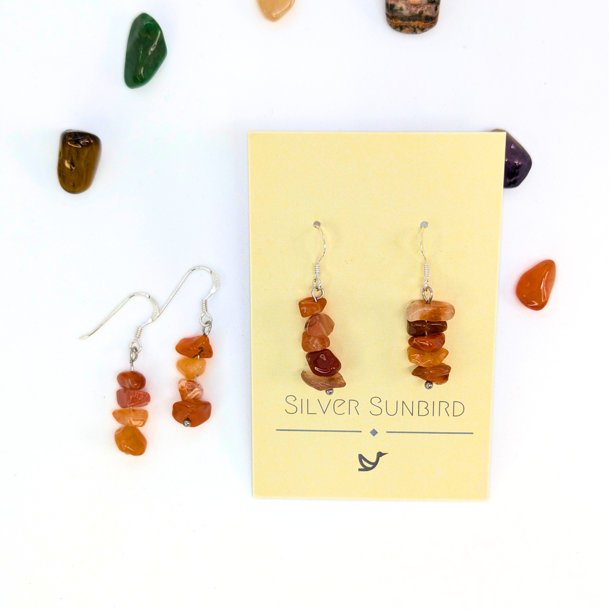 Carnelian Agate Earrings by Silver Sunbird: sterling silver dangle earrings with stacked orange gemstones, displayed on a branded card. Polished multicolored gemstones are scattered on a white background.